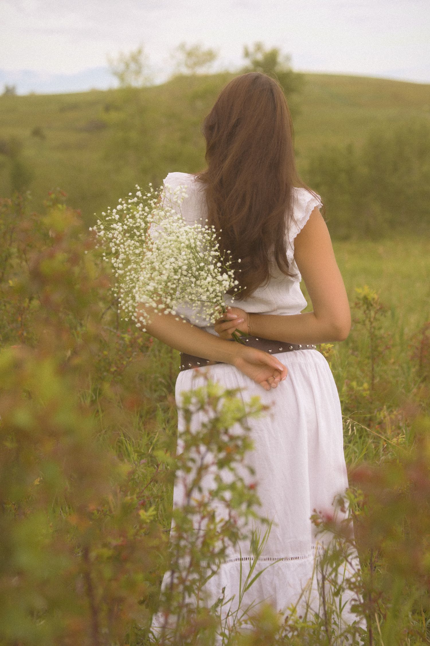 A person in a white summer dress poses with flowers in a scenic hillside meadow.