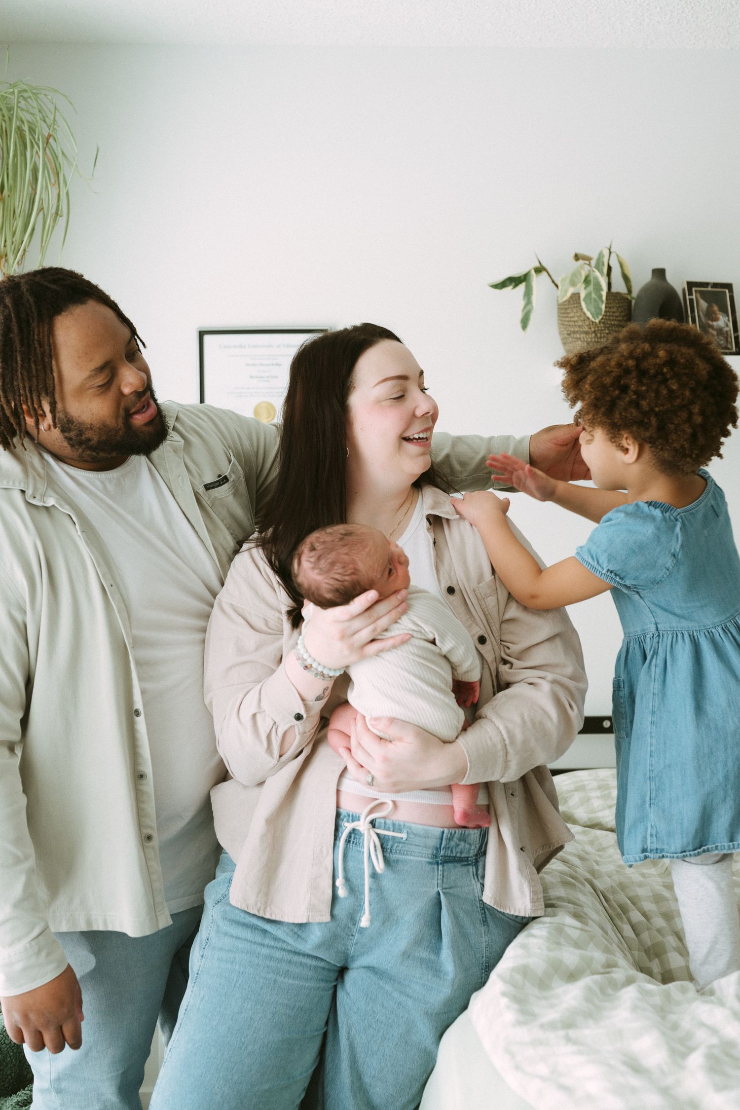 Happy interracial family with parents and two children, including a newborn baby, sharing a joyful moment in a bright bedroom.
