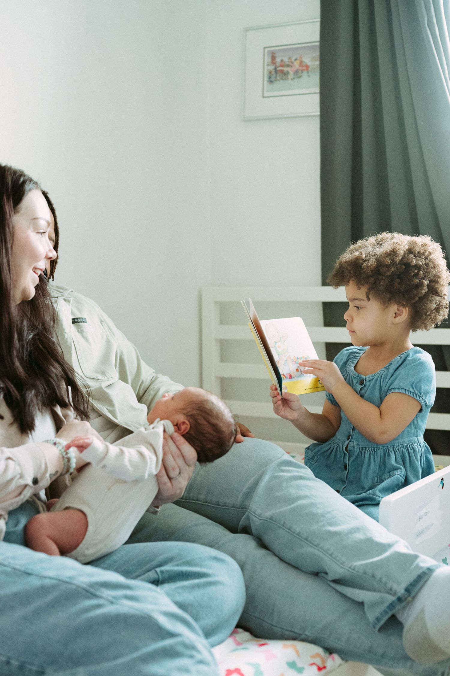 A happy mother holds her newborn while a curly-haired toddler shows her a colorful book on a bed.