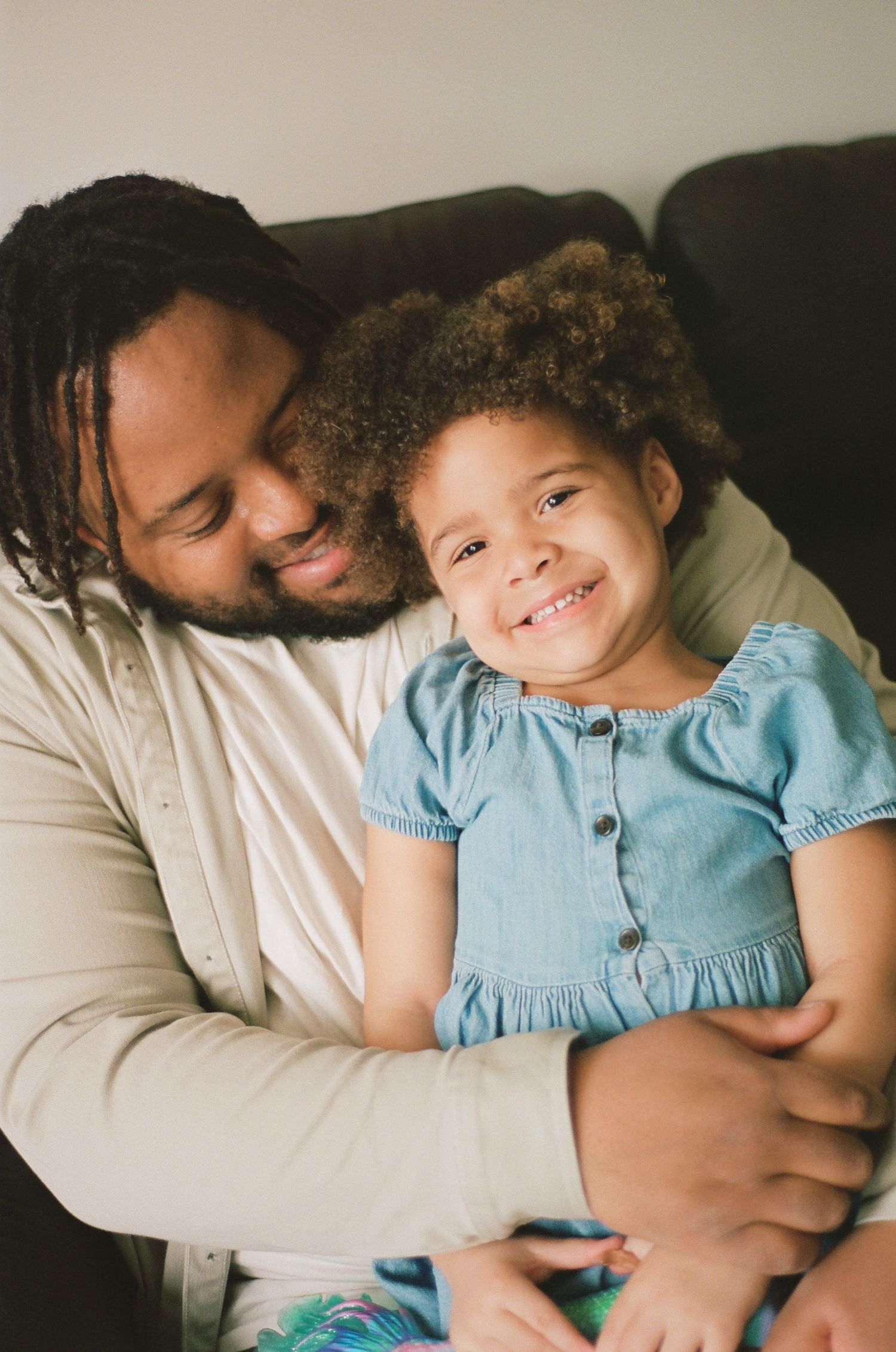 A father lovingly embraces his smiling young daughter in a blue dress, sharing a warm moment together at home.