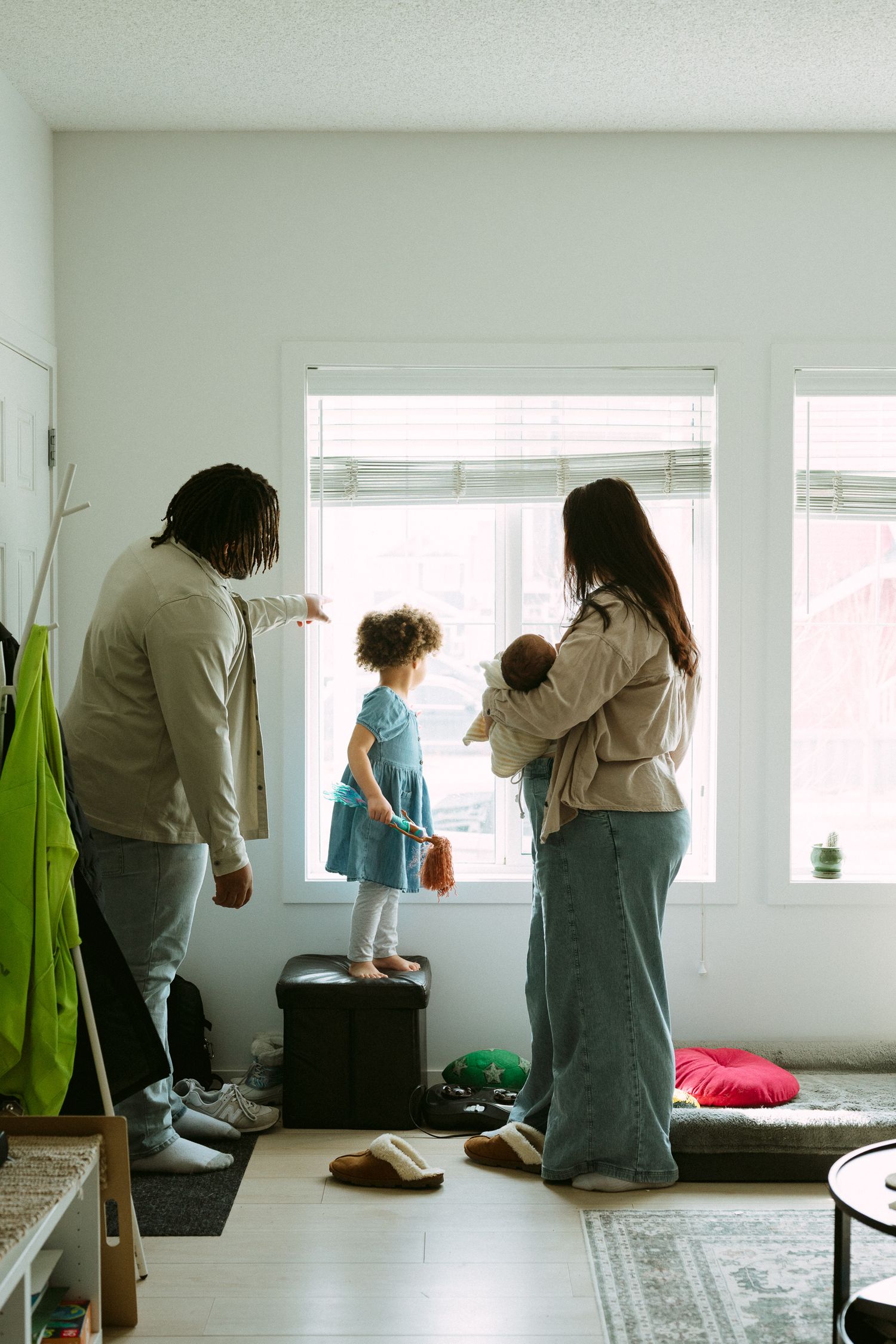 A family of four stands by a bright window, with parents and two young children looking outside in a cozy home entryway.