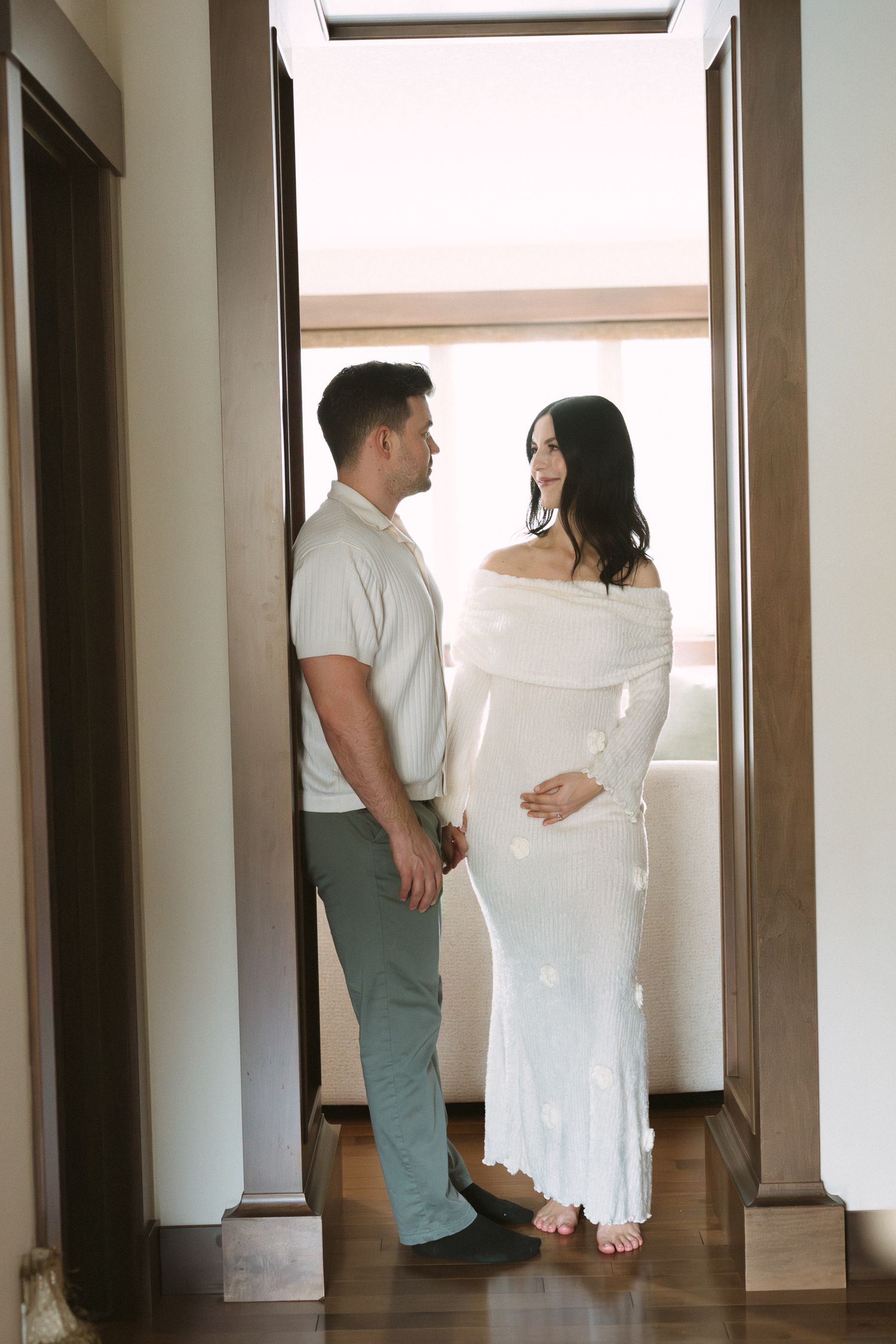 Expecting couple facing each other in a bright doorway, woman in white off-shoulder maternity dress.