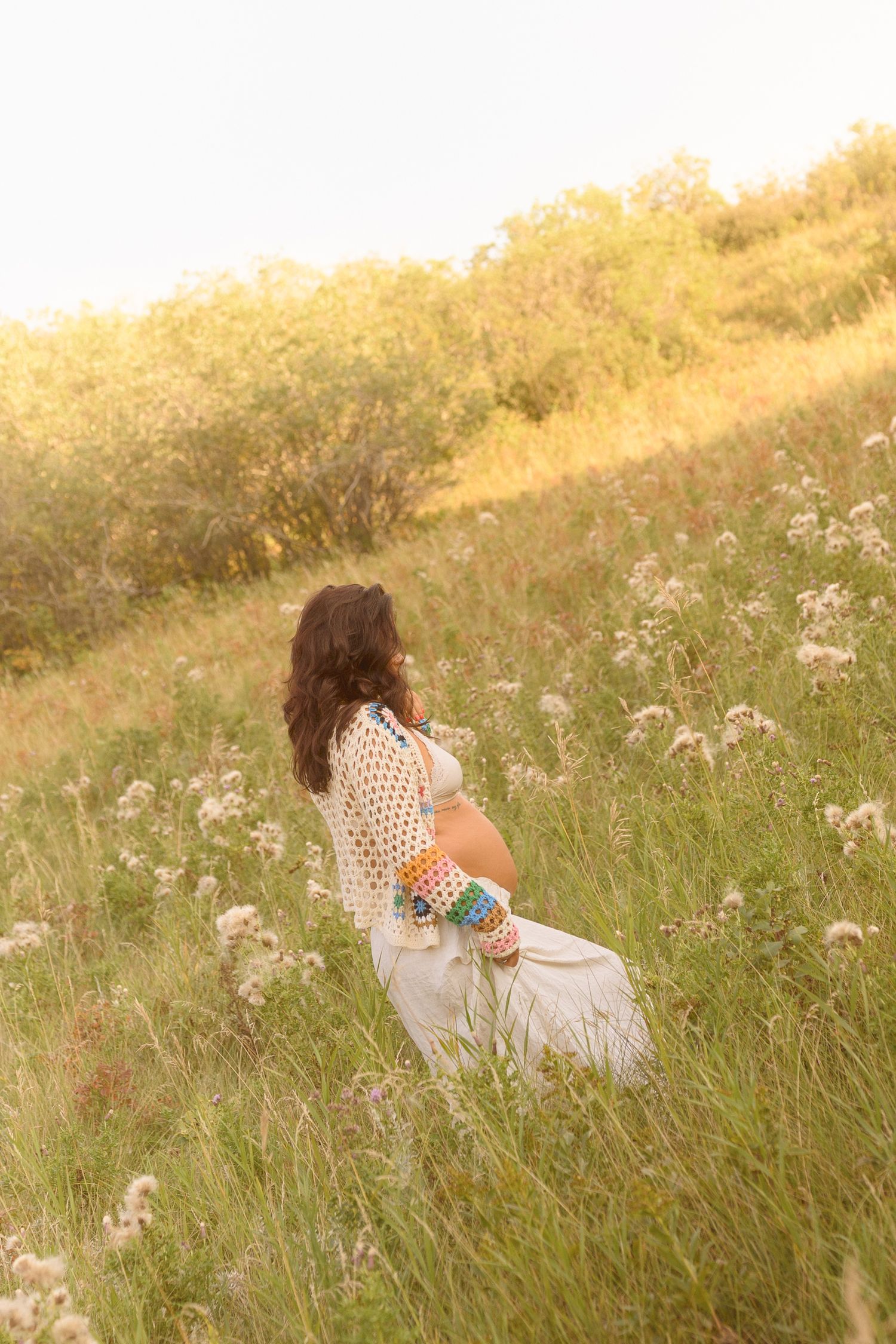 An expectant mother poses gracefully among tall grasses in a sunlit meadow wearing white.