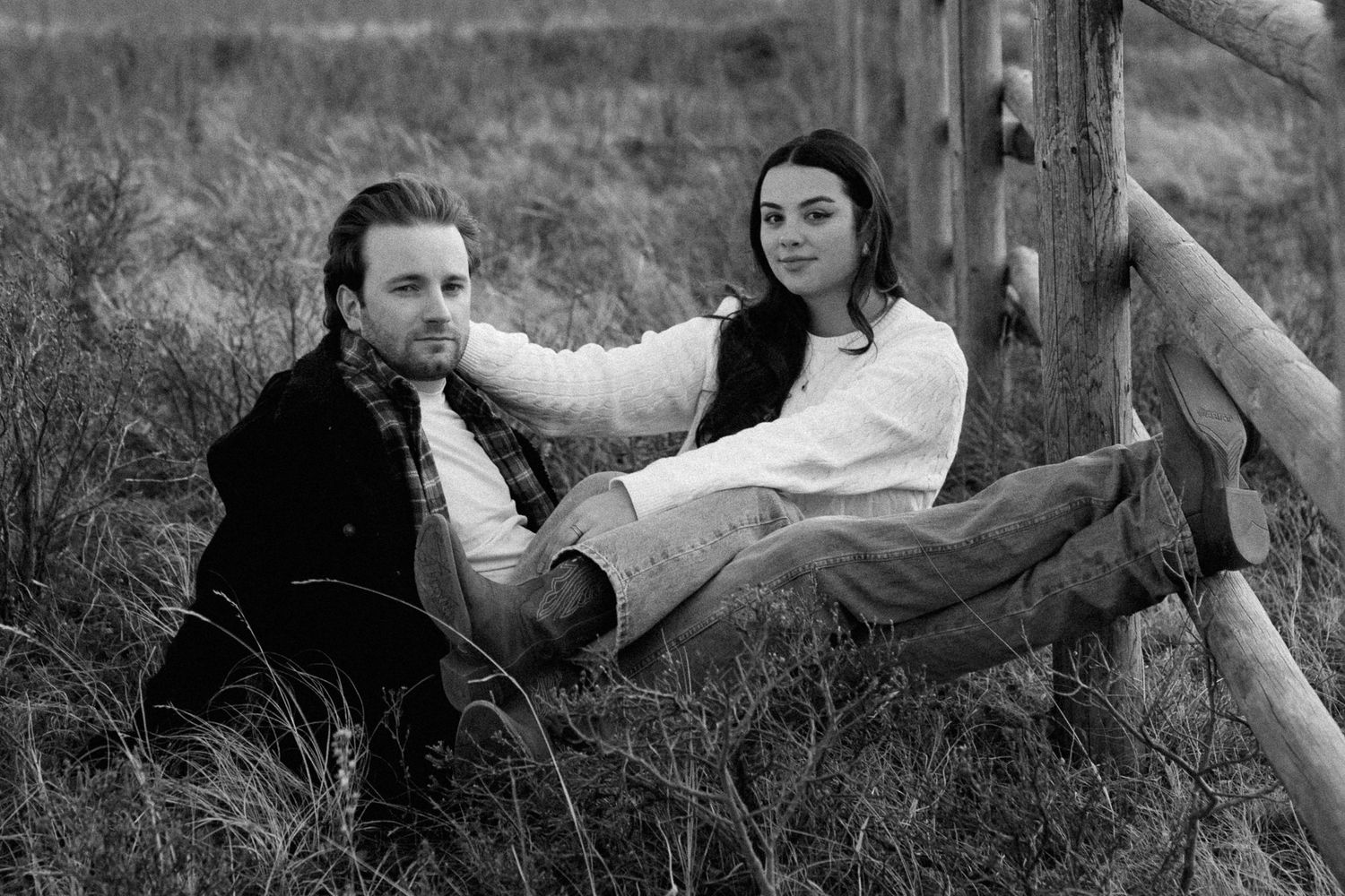 A couple relaxes together in black and white against a rustic wooden fence in a grassy field.