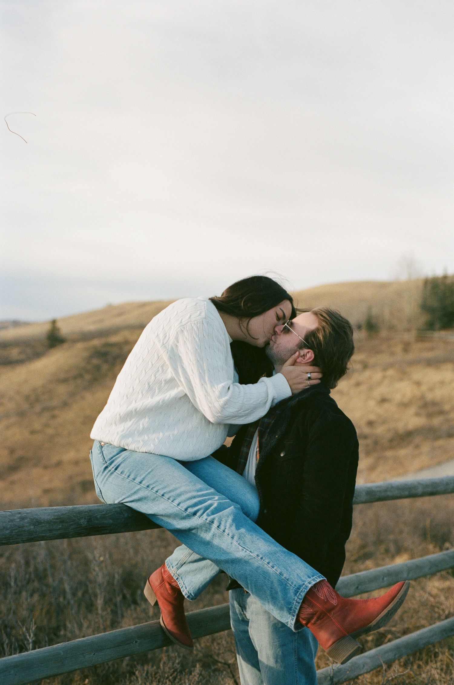A couple in casual attire embraces while leaning on a wooden fence with rolling hills in the background.