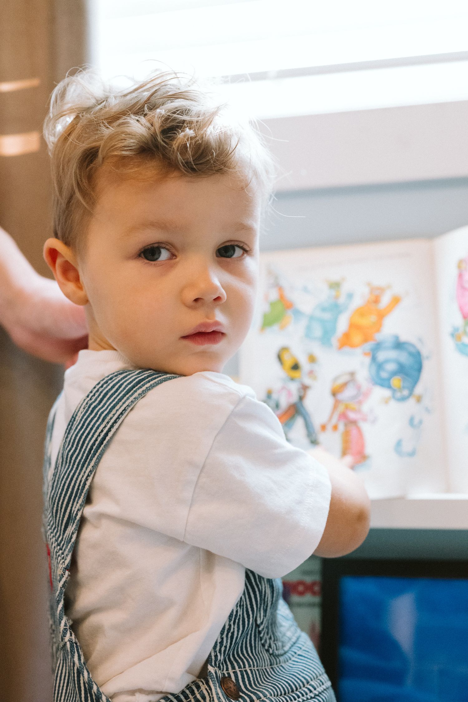 A toddler in a striped shirt and white sleeves looks to the side while colorful artwork decorates the wall behind.