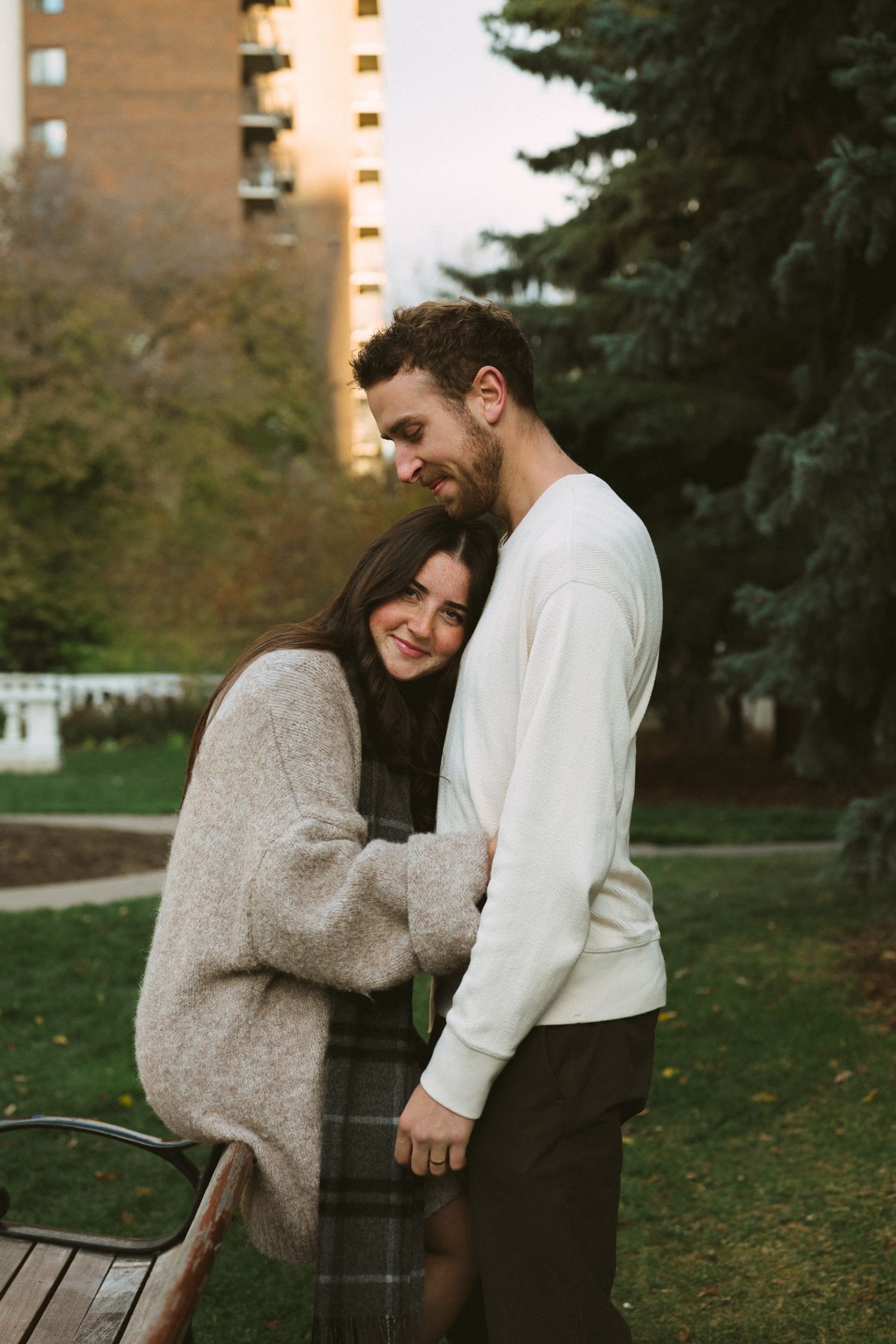 A couple shares a tender moment together outdoors on an autumn day, laughing and embracing in casual attire.