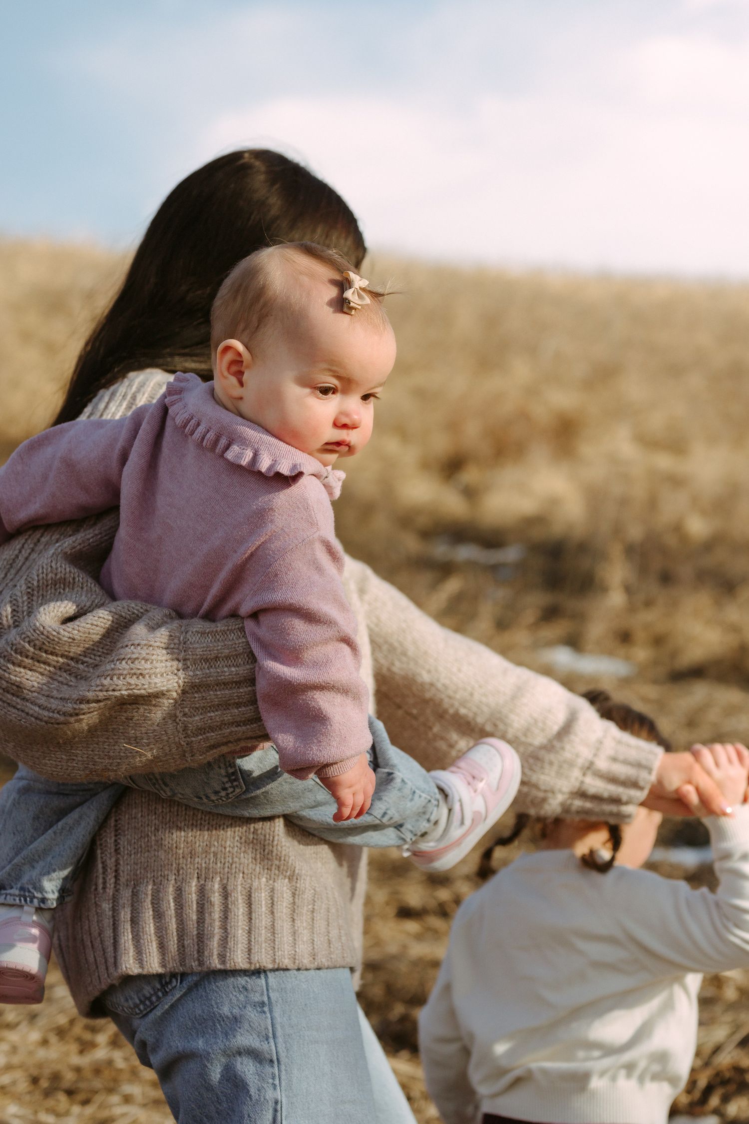 A baby wearing a pink sweater is carried on someone's back while outdoors in a field during golden hour.