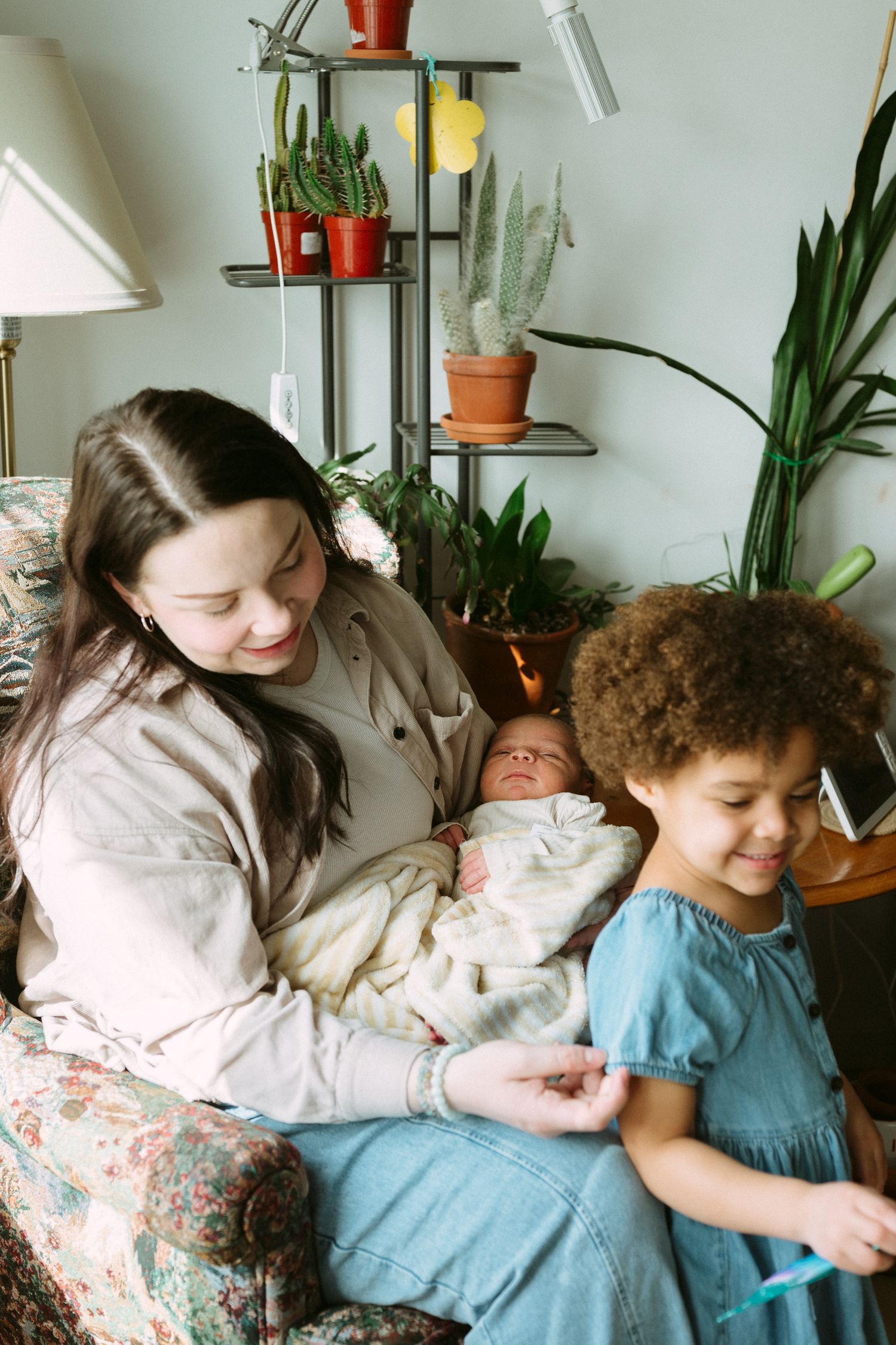 A smiling mother sits in a floral armchair holding a newborn while a young curly-haired girl stands beside her among houseplants.