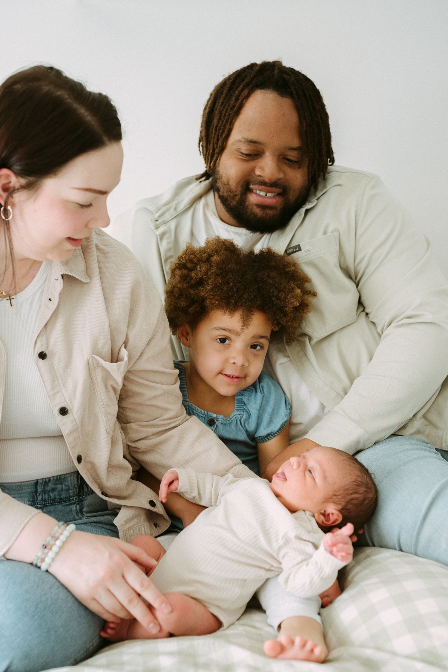 A happy interracial family of four sits together, parents smiling at their newborn baby while a young child looks on.
