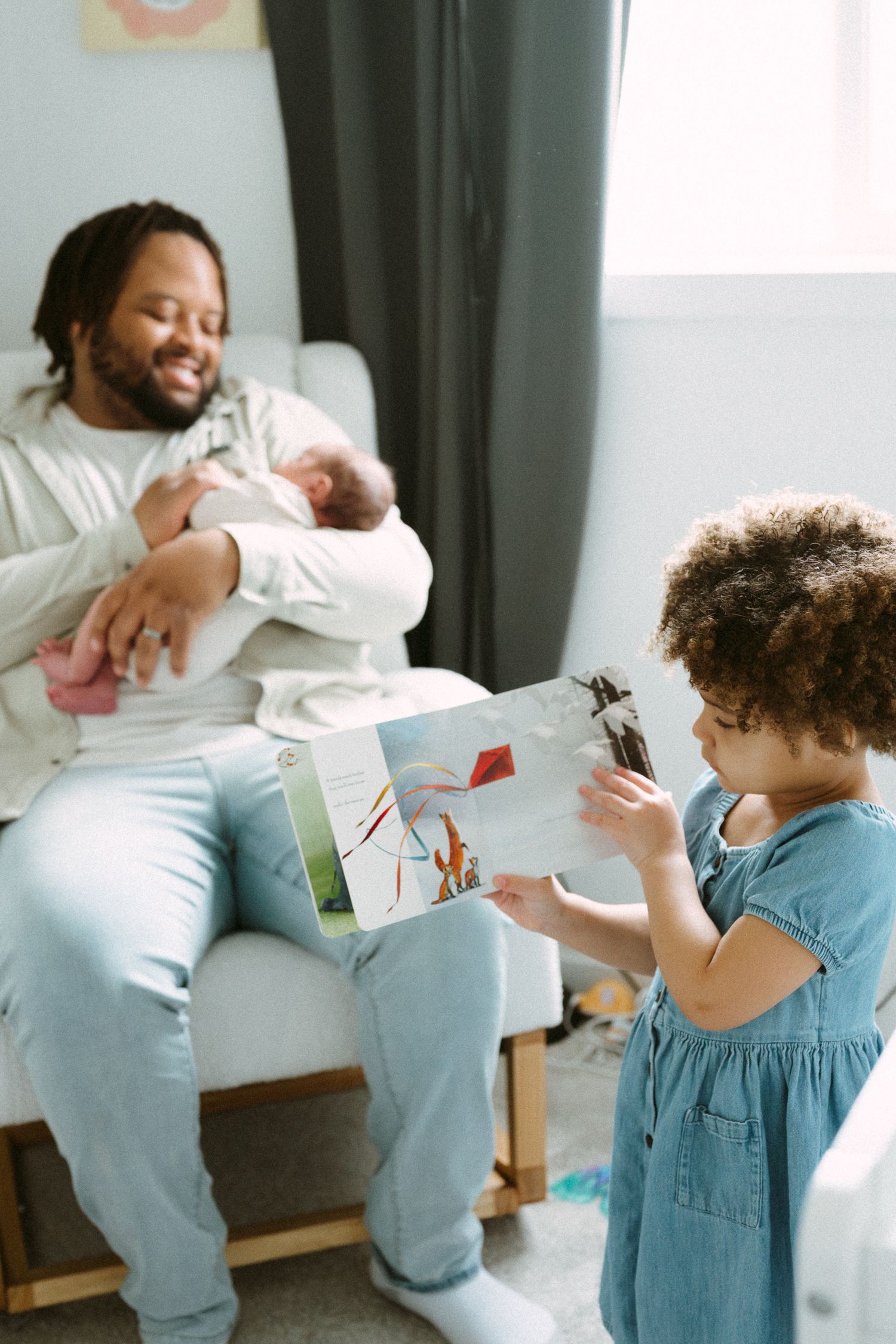 Smiling father holds newborn baby while young curly-haired girl shows him a picture book in nursery.