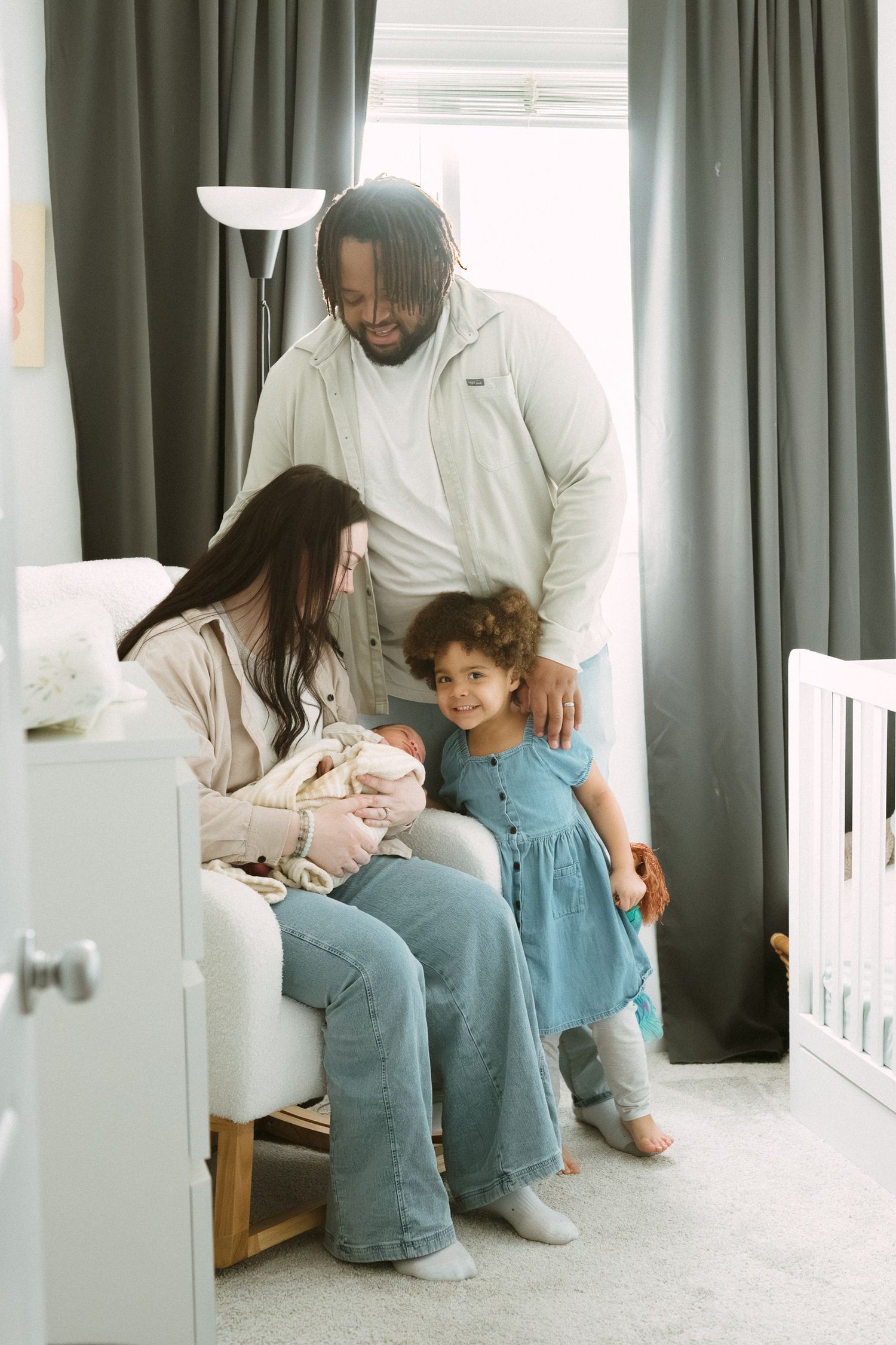Interracial family with mother, father, toddler girl, and newborn baby gathered in a nursery room.