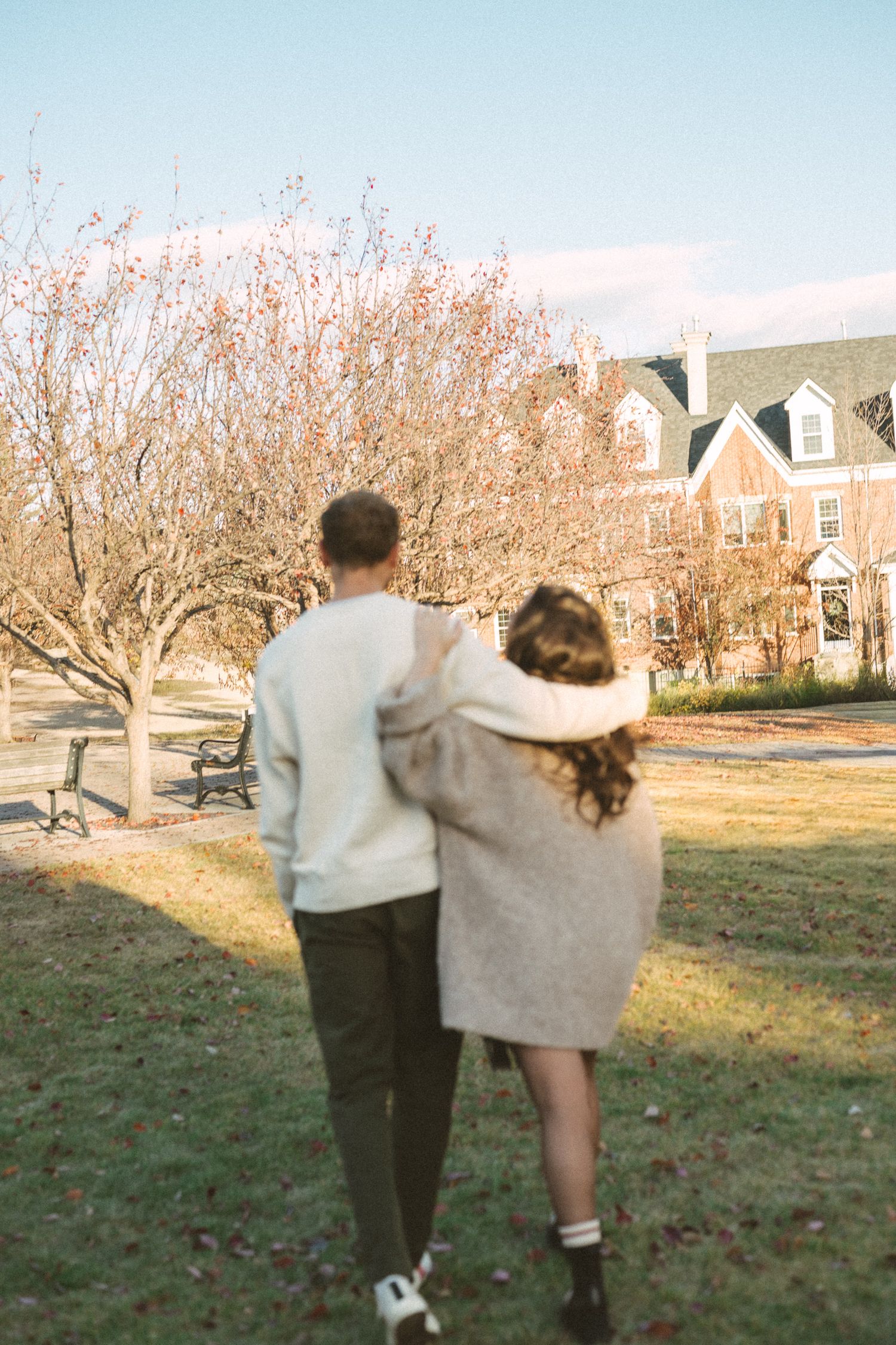 A couple walking together on a grassy lawn during sunset with autumn trees and houses in the background.