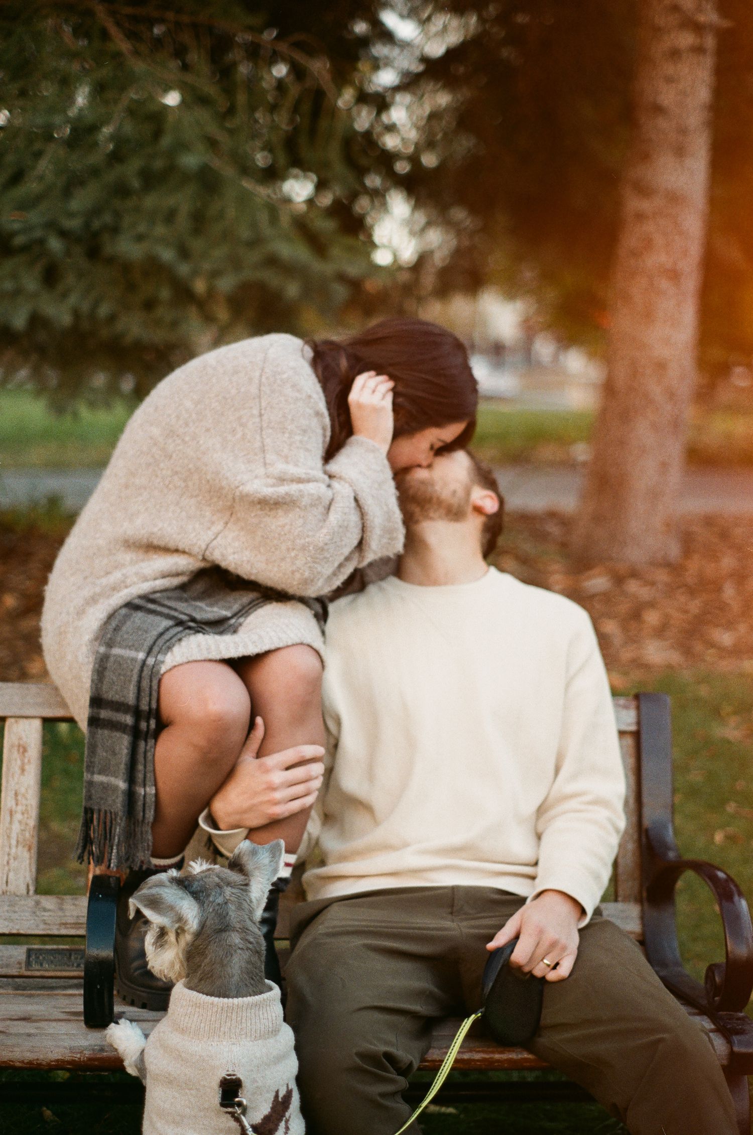 A couple shares an intimate moment on a park bench at sunset while their dog sits beside them.