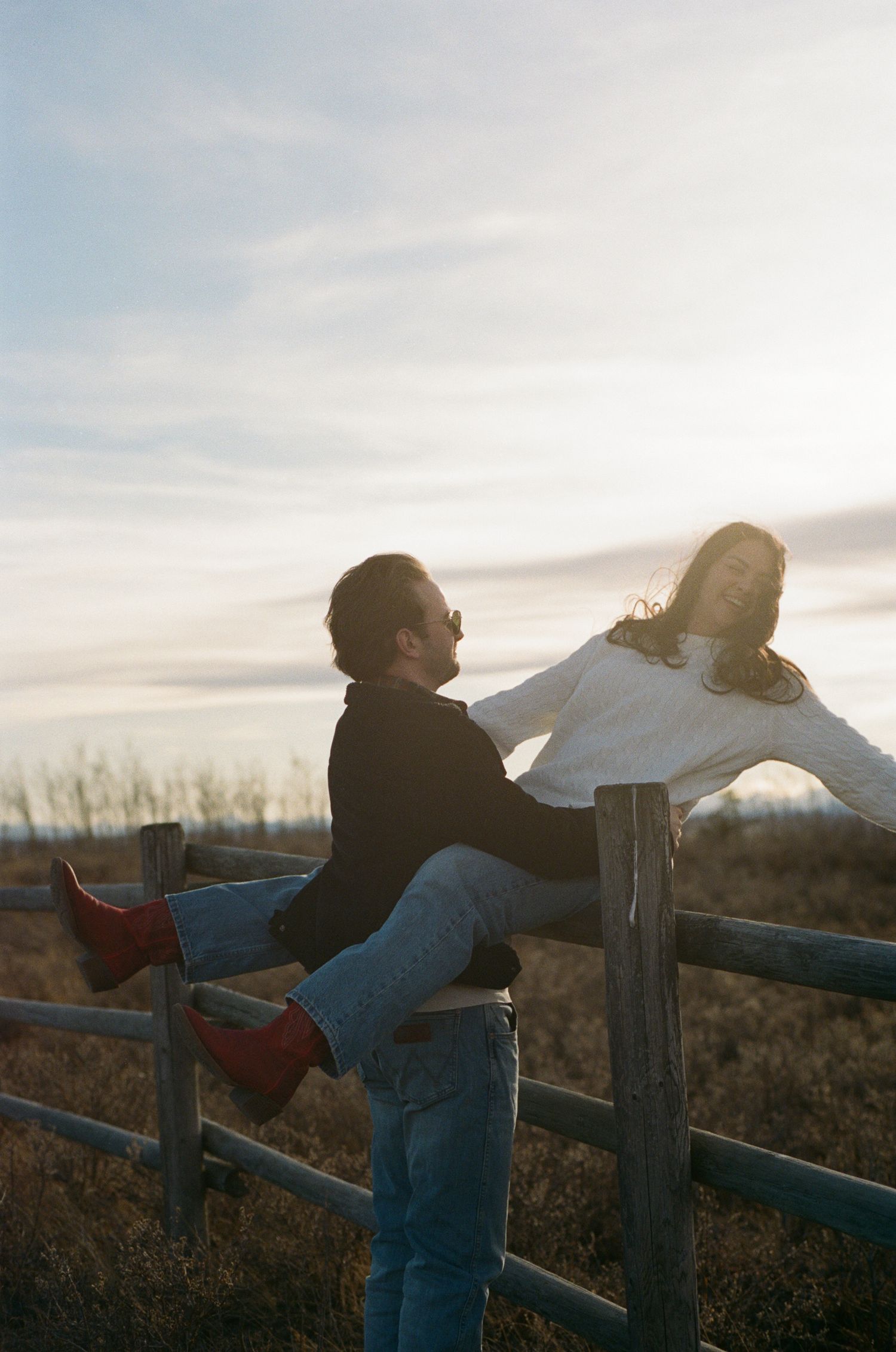 Couple playfully interacting by a wooden fence at sunset in a rural field.