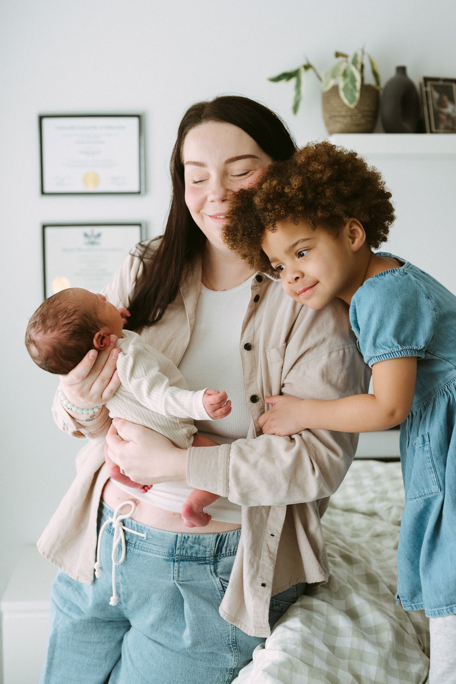A smiling mother holds her newborn baby while her young curly-haired daughter hugs her in a bright bedroom.