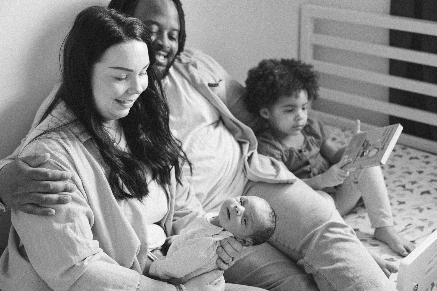 Black and white photo of a happy interracial family relaxing on a bed with a newborn baby and toddler.