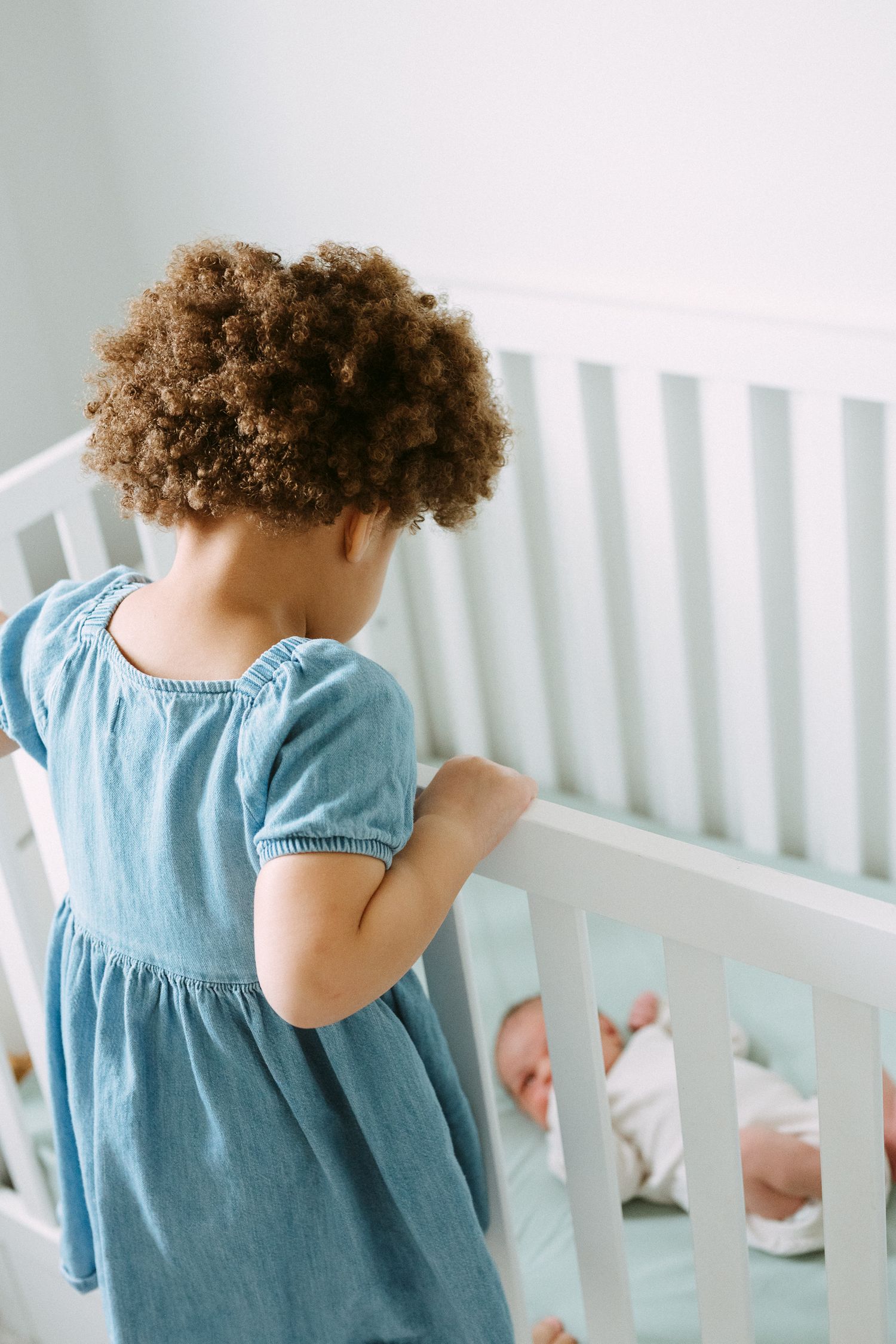 Curly-haired toddler in blue dress standing at white crib looking at newborn baby sibling.