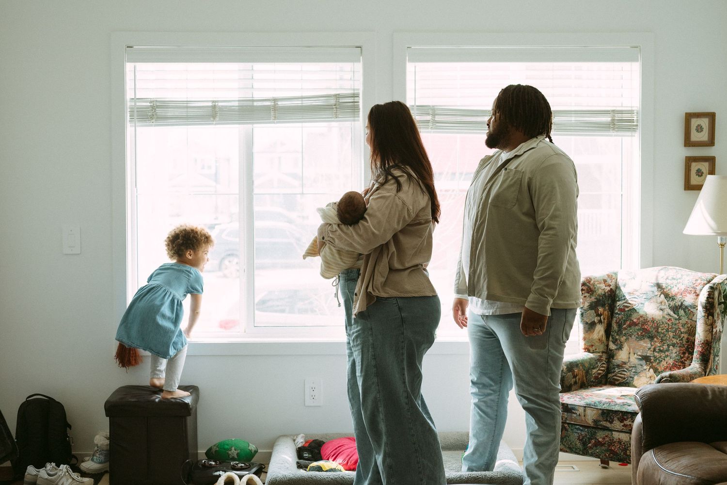 Family of four standing together looking out bright living room windows, parents holding newborn baby with toddler nearby.