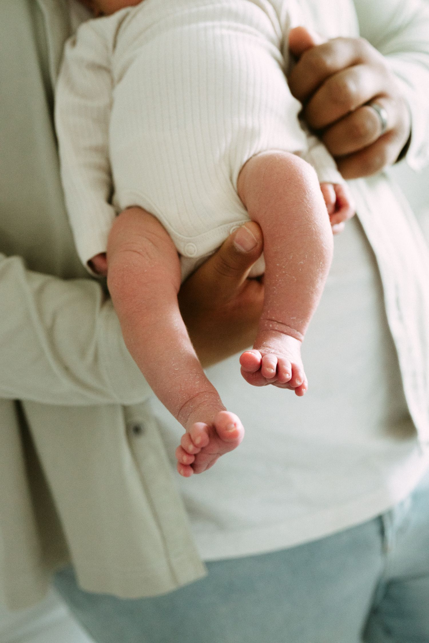 Close-up of newborn baby in white onesie being held by a parent with a wedding ring.
