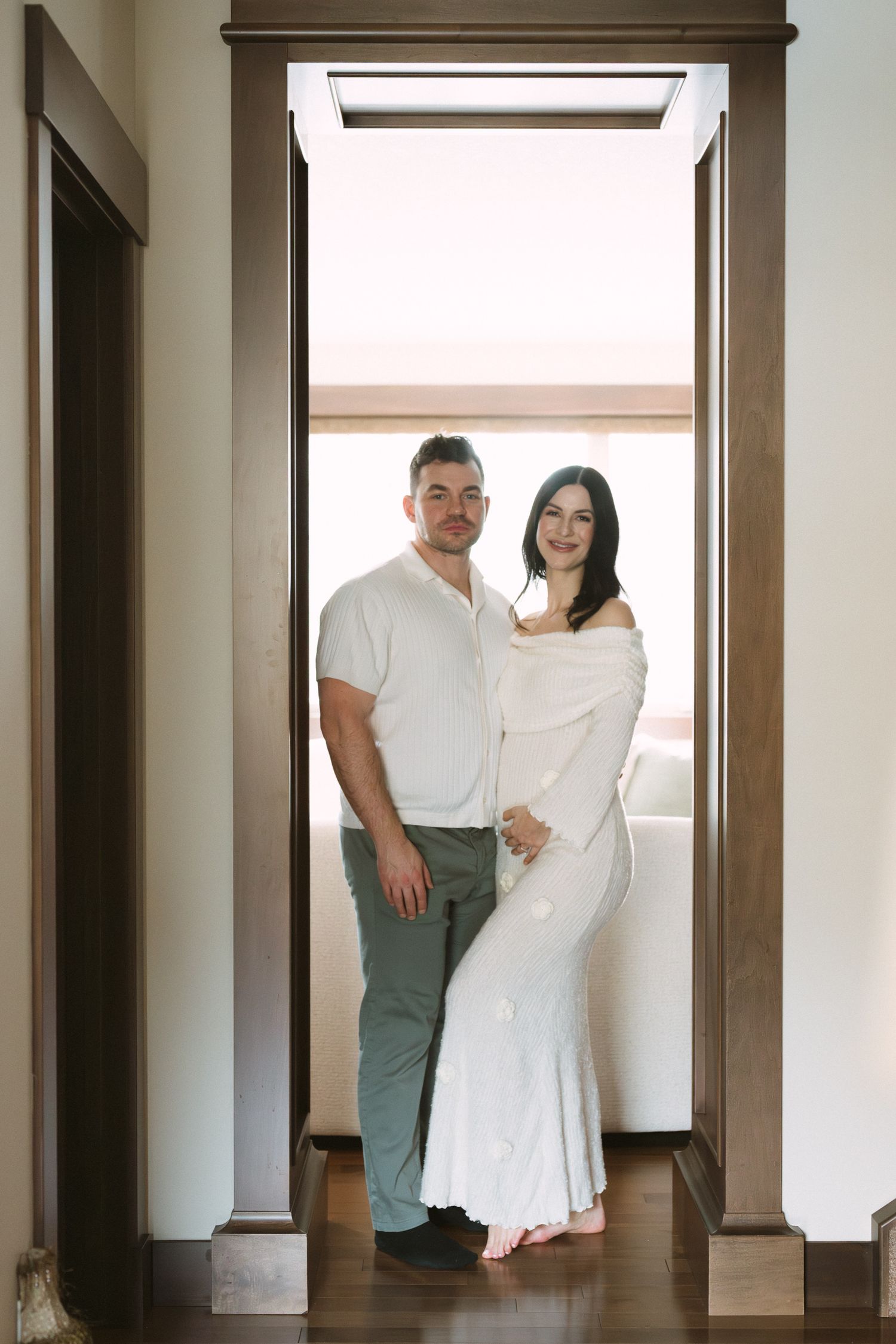 Couple posing in wooden doorframe, pregnant woman in white off-shoulder maternity dress standing with partner.
