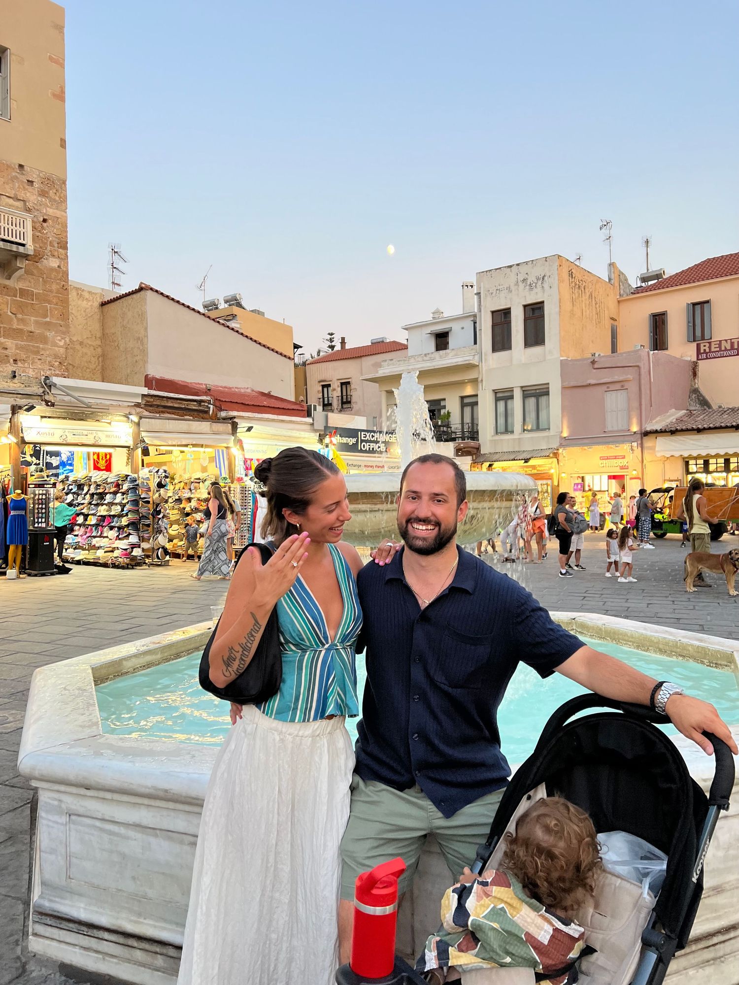 A couple enjoys an evening at a lively town square with shops and a fountain in the background.