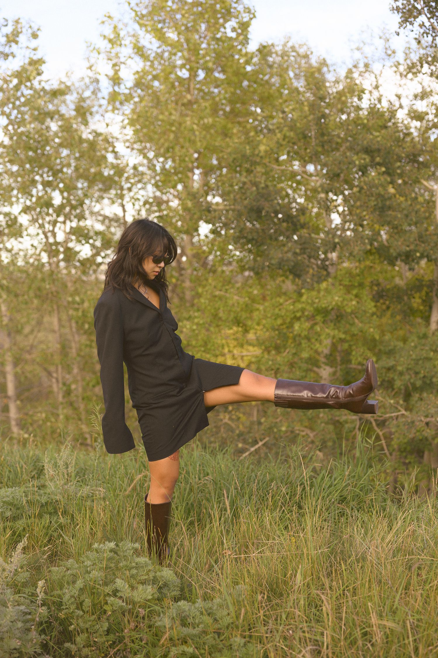 Person in black dress and boots playfully kicks leg up while posing in tall grass.
