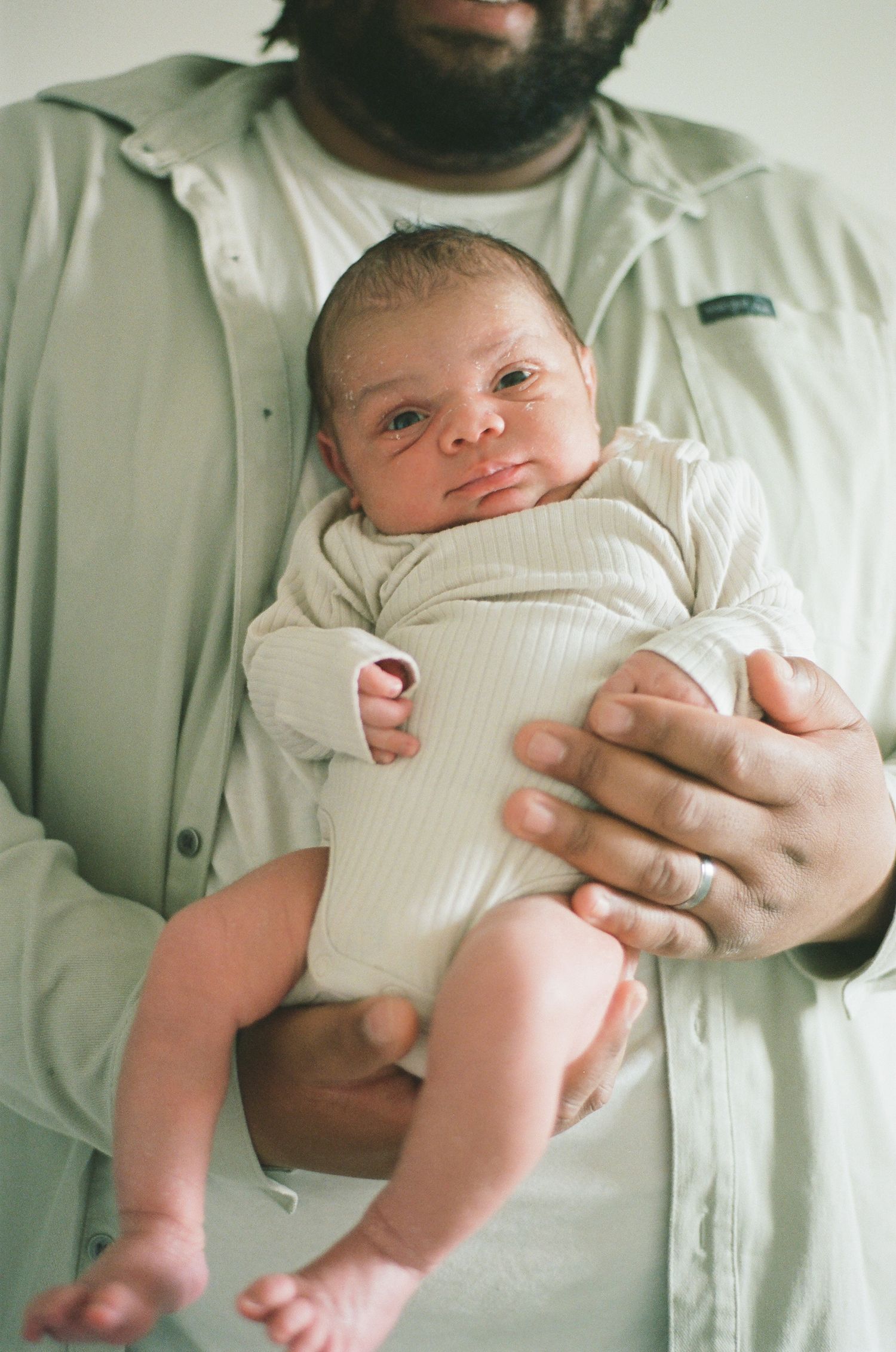 Father in sage green jacket cradling alert newborn baby in white ribbed onesie against his chest.