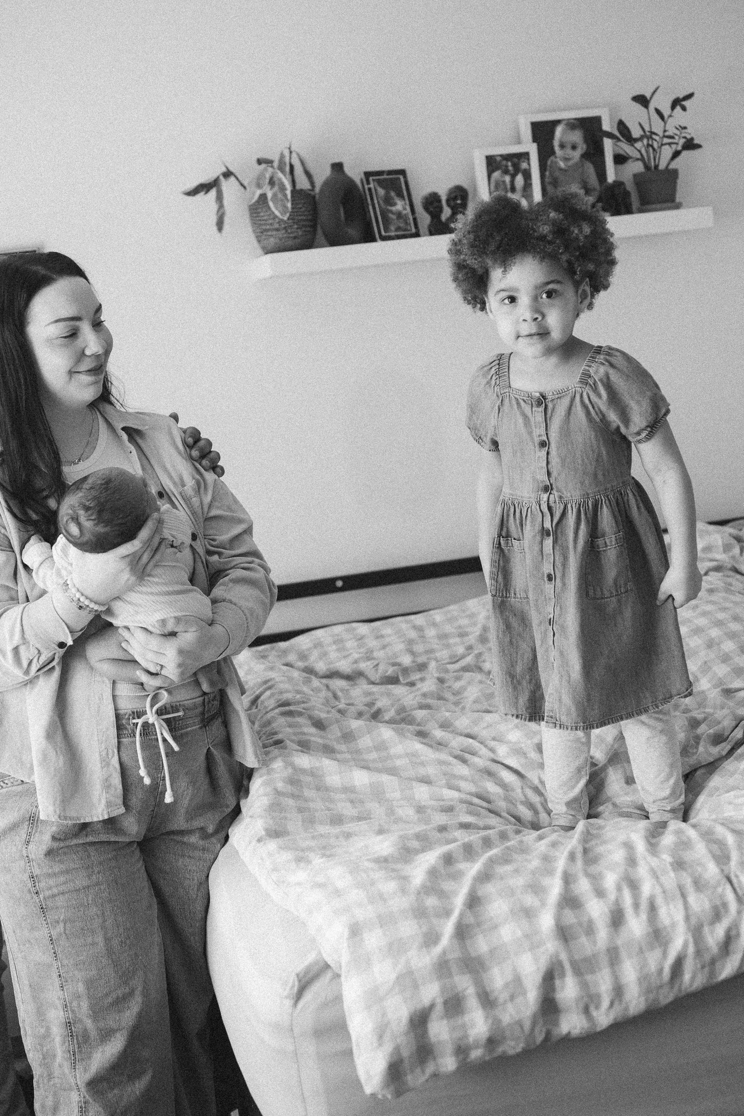 Black and white photo of a mother holding a newborn while a curly-haired toddler stands on a bed.