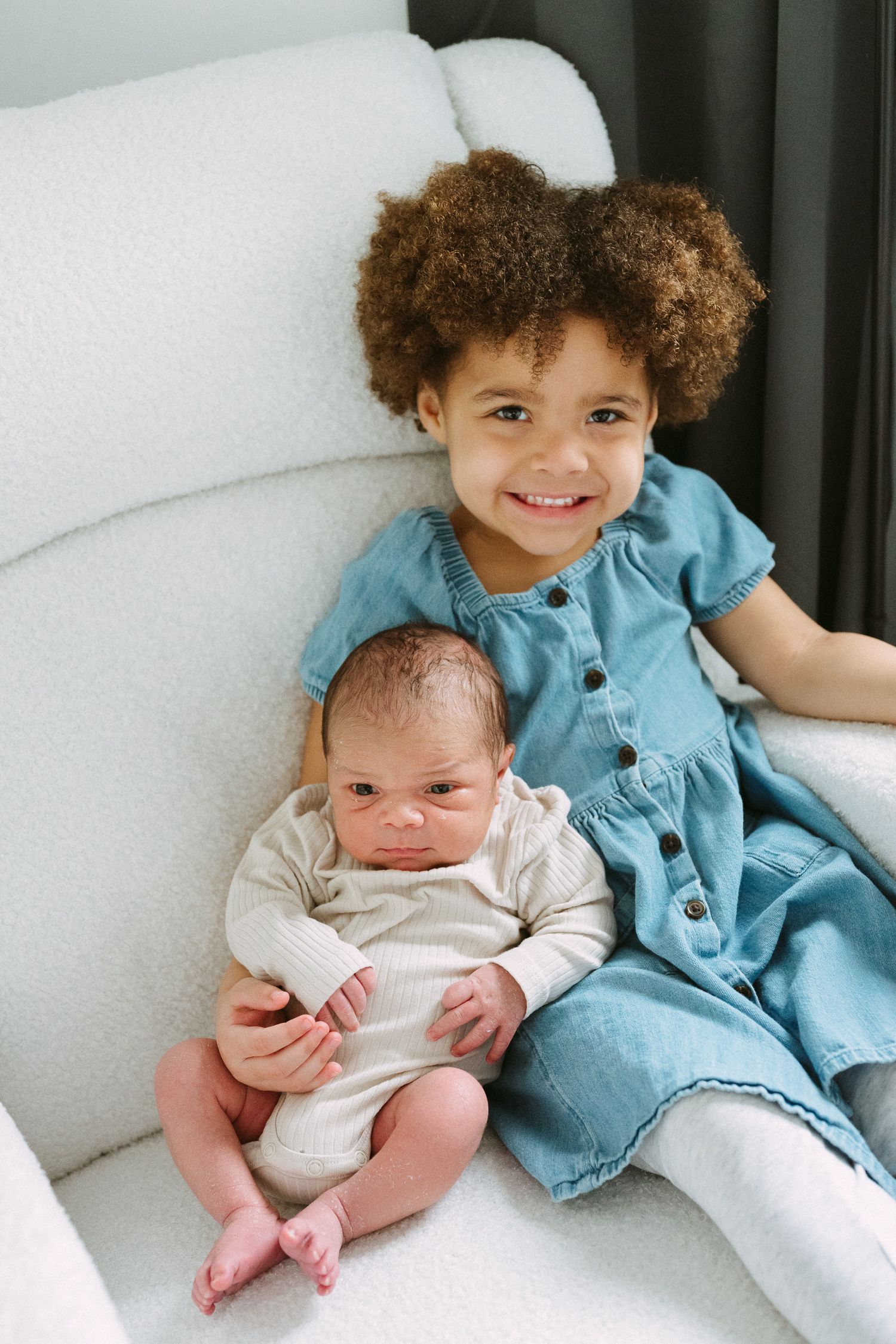 Happy curly-haired girl in denim dress smiles while holding newborn baby sibling on white armchair.