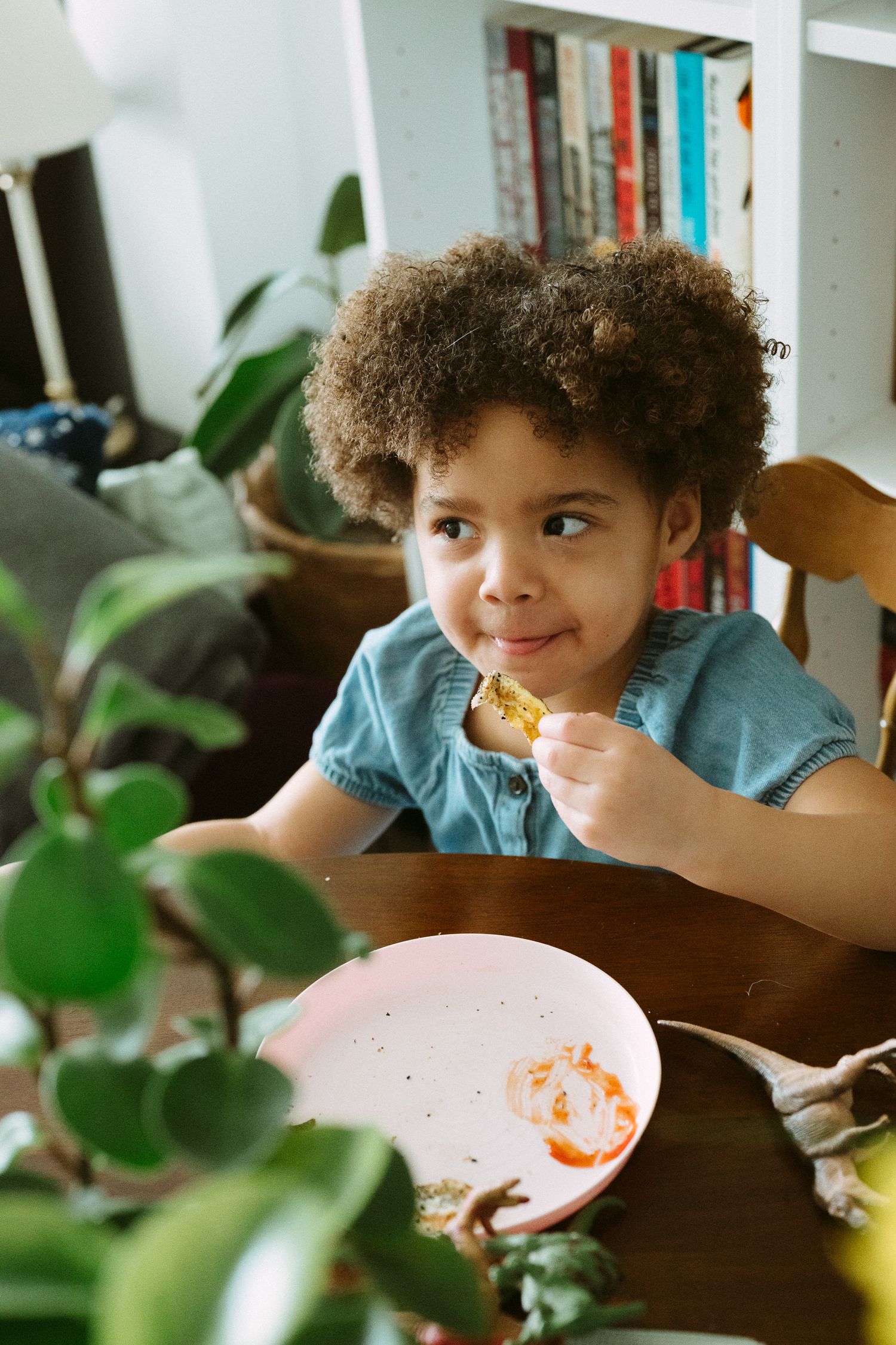 A curly-haired young girl in a blue dress eats at a wooden table with a pink plate, surrounded by houseplants.