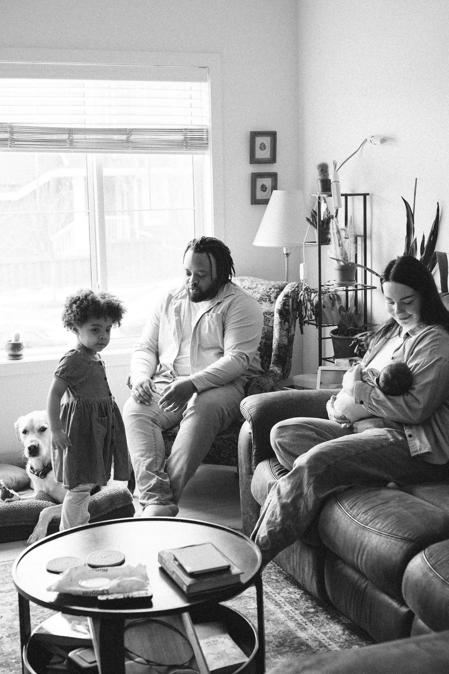A family with a toddler and newborn baby relaxes together in a cozy living room in black and white.