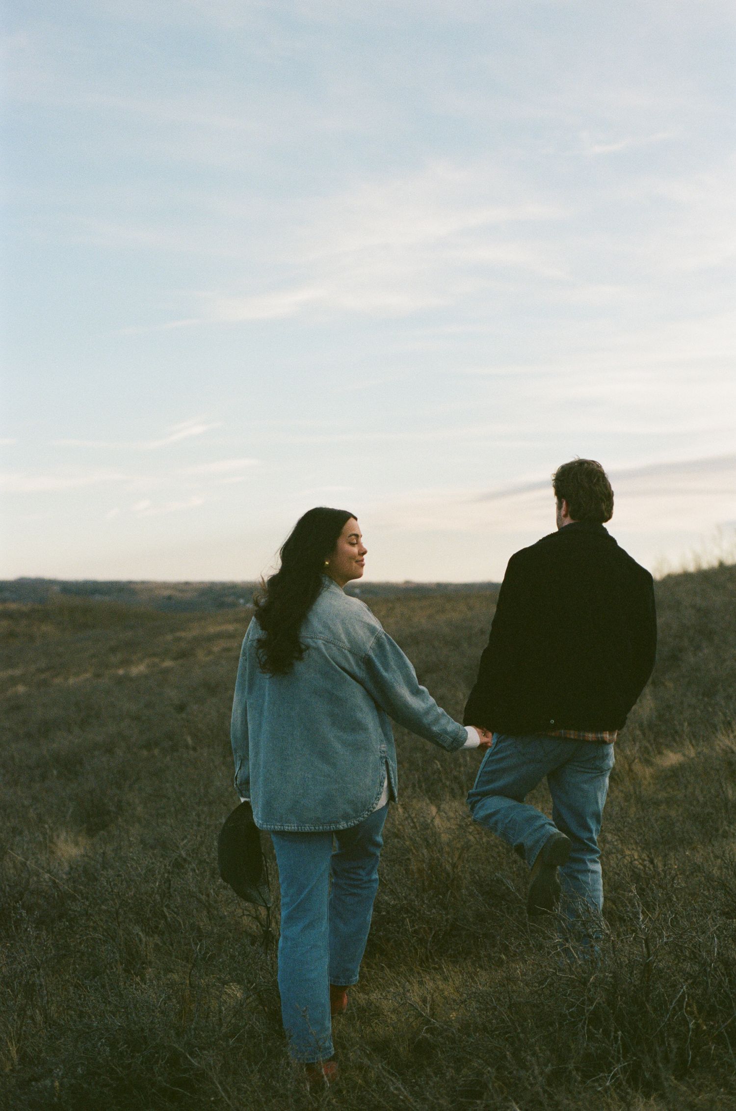 Two people in denim outfits walk together across a grassy hillside at dusk.