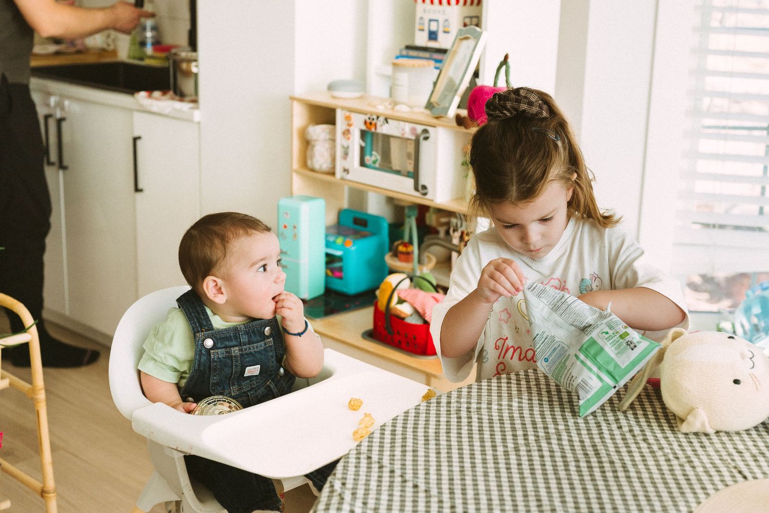 Mealtime scene with toddler in highchair and caregiver at kitchen table.