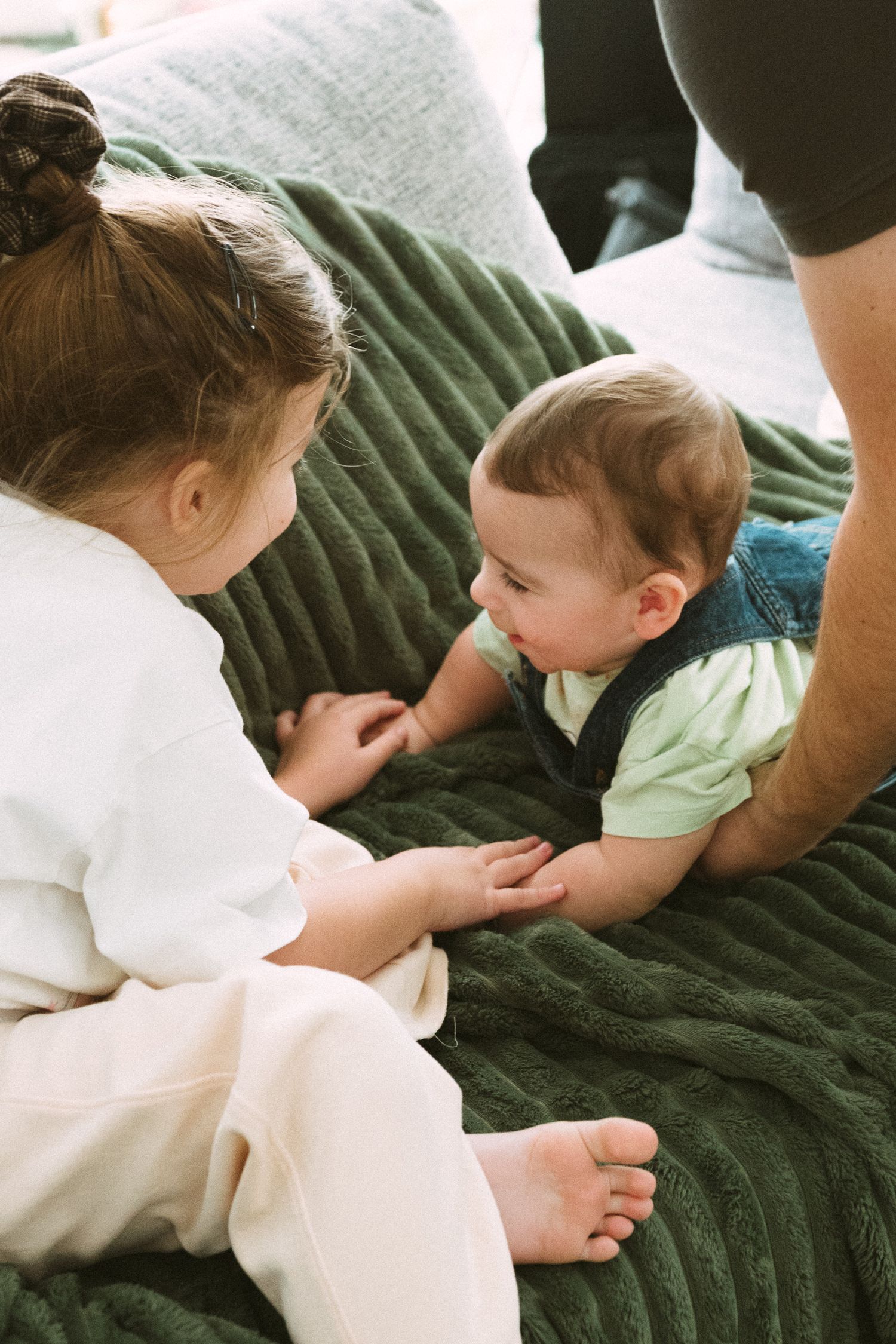 Two young children sit together on a dark green textured blanket while interacting with each other.