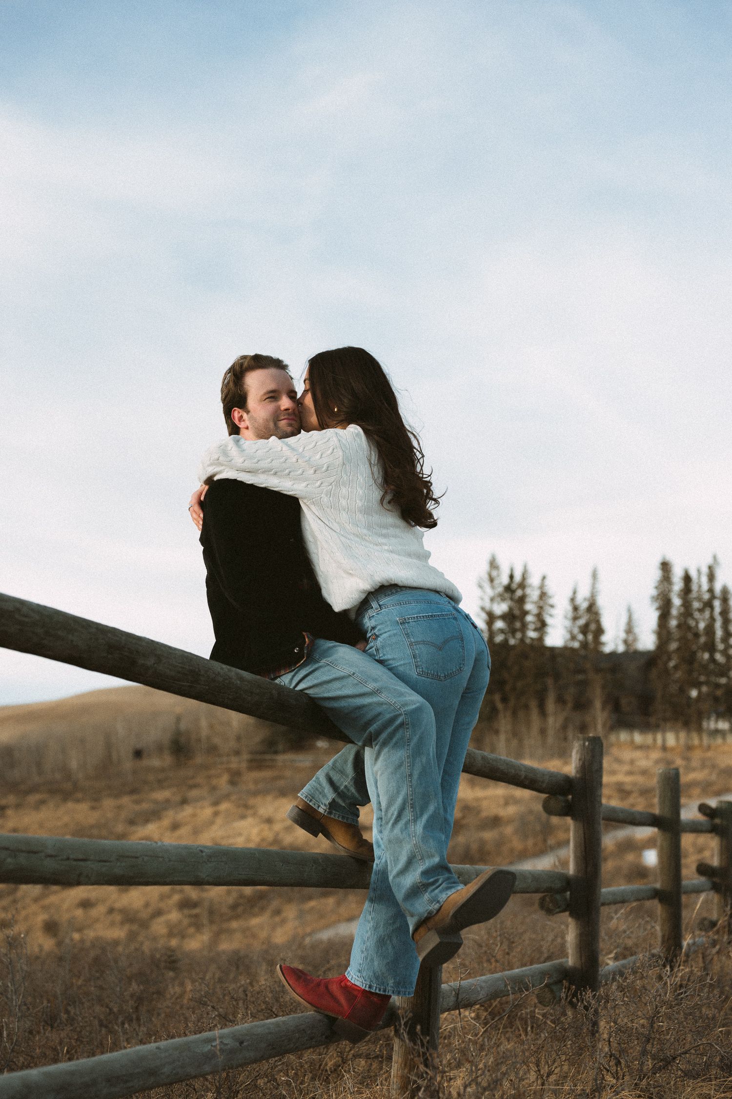 A couple sits intimately on a wooden fence overlooking a rural landscape with hills and trees at sunset.