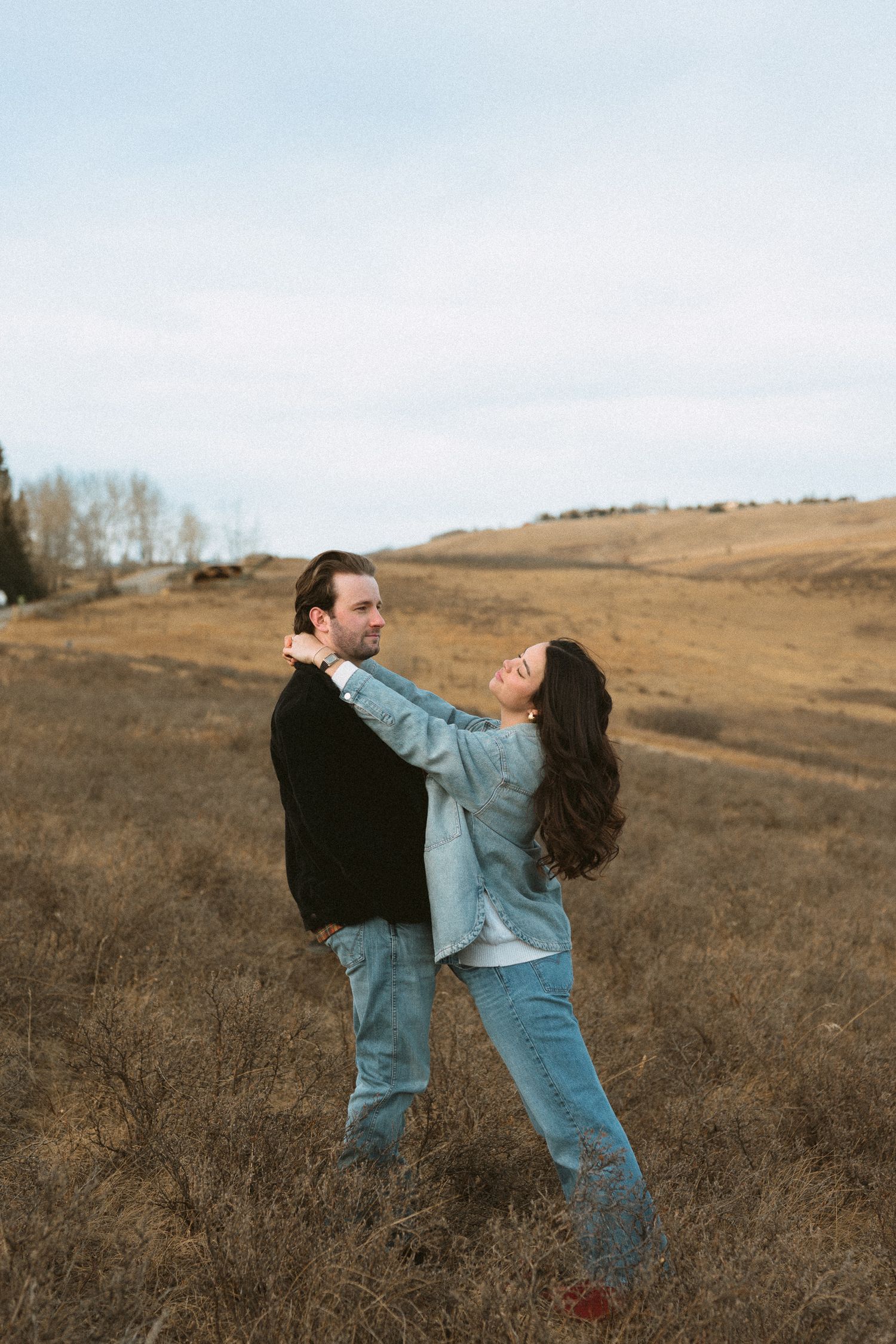 A couple embraces playfully in a rustic field with brown grass and rolling hills against an overcast sky.