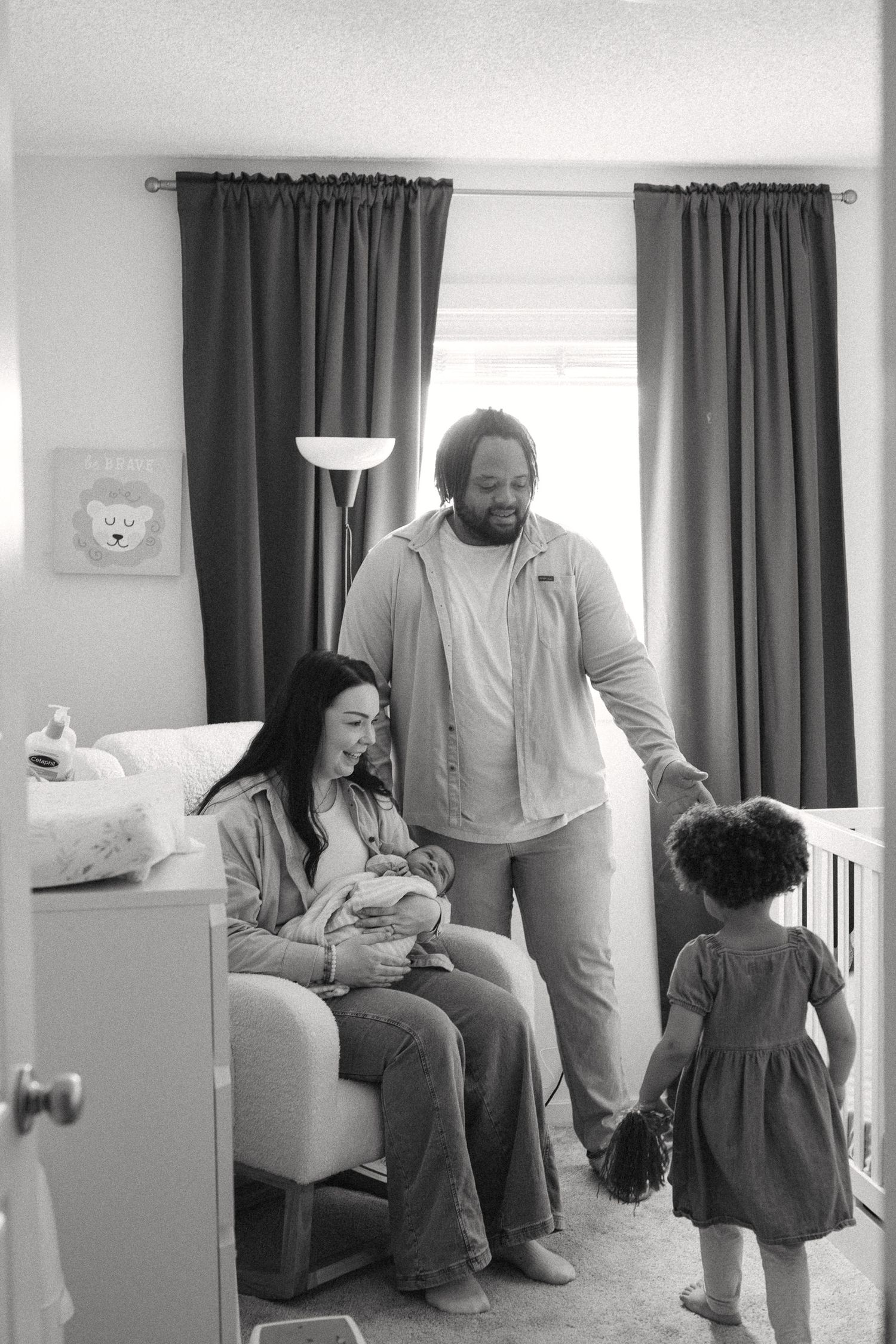 Black and white photo of a family with a newborn baby in a nursery, mother seated holding infant while father and toddler stand nearby.