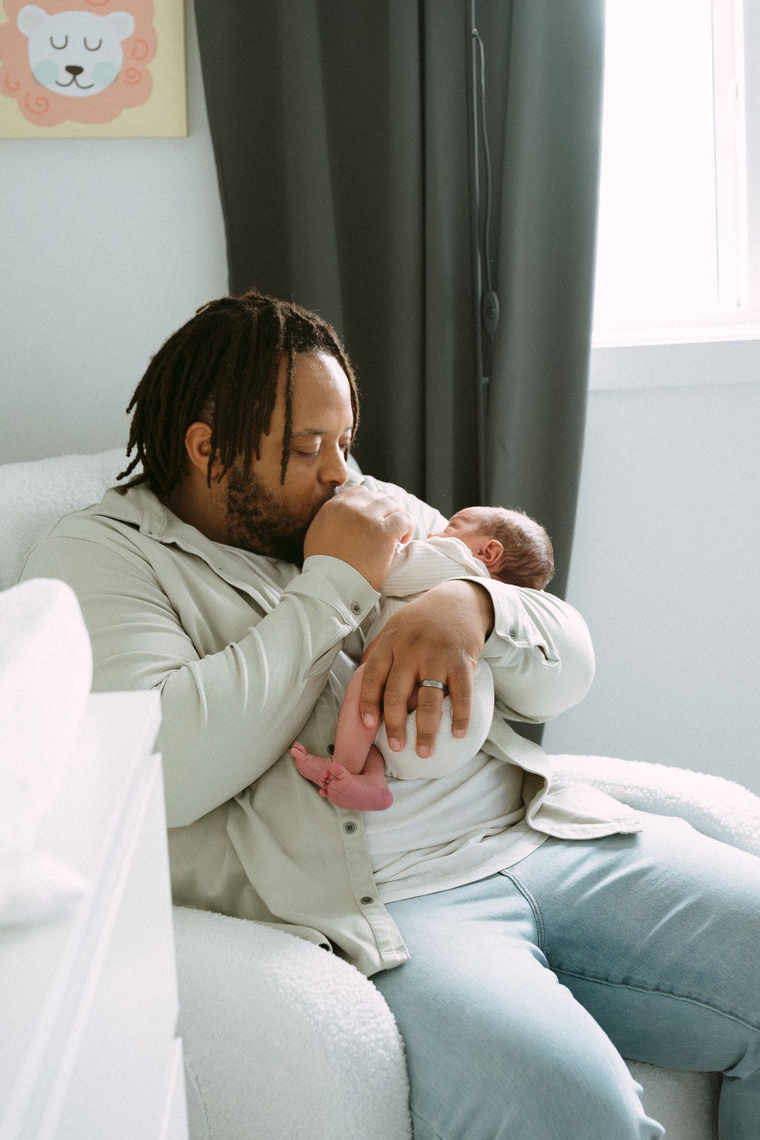 A father with dreadlocks sits in a nursery chair, gently kissing his sleeping newborn baby.