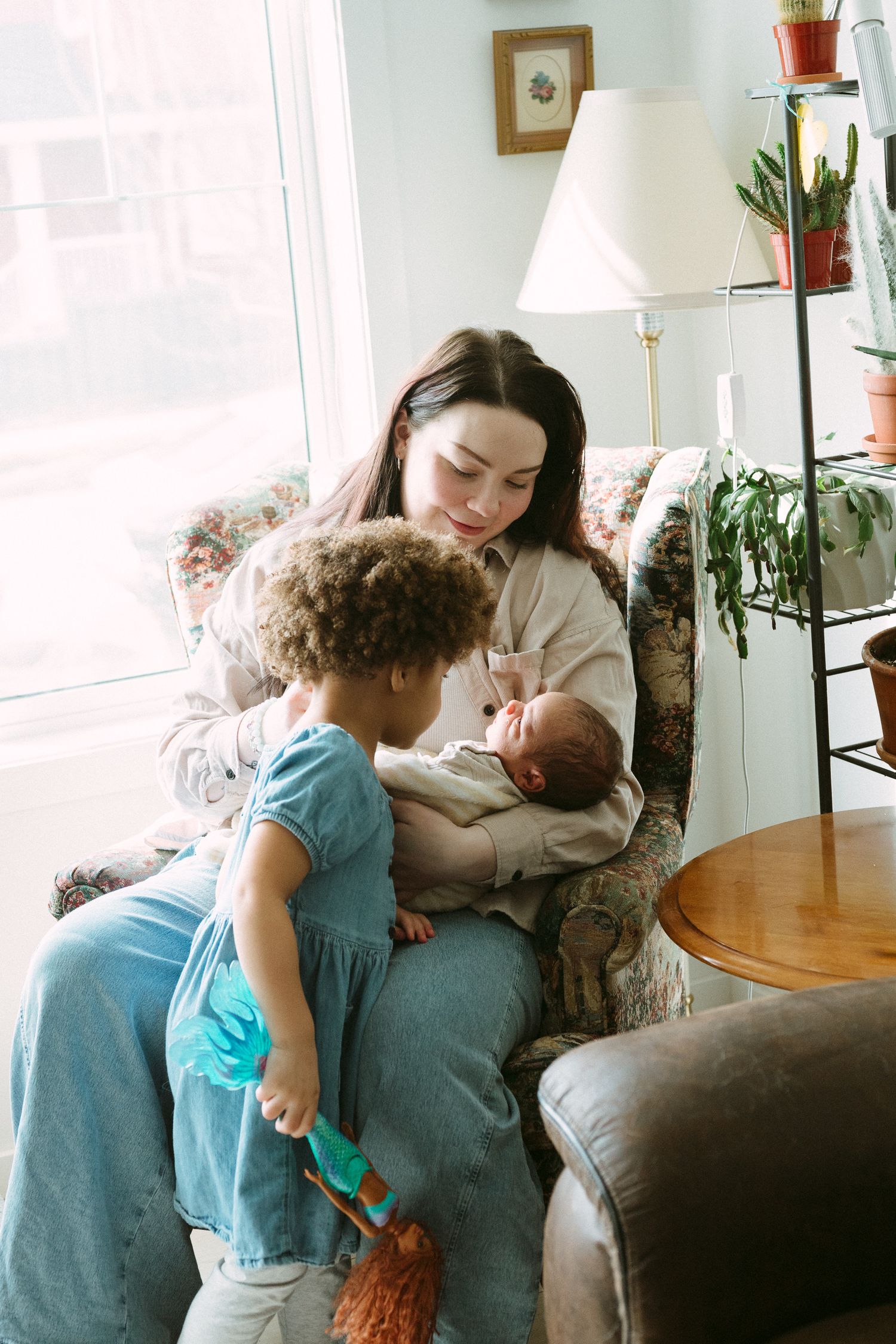 A mother sits in a floral armchair holding a newborn while a toddler in a blue dress looks on in a cozy living room.