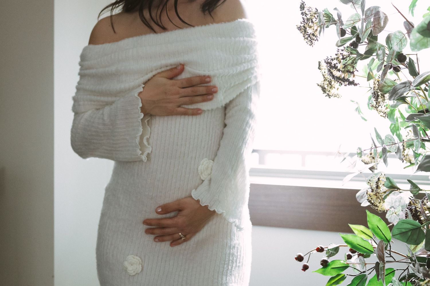 Close-up of a pregnant woman in a white knit off-shoulder dress cradling her baby bump.
