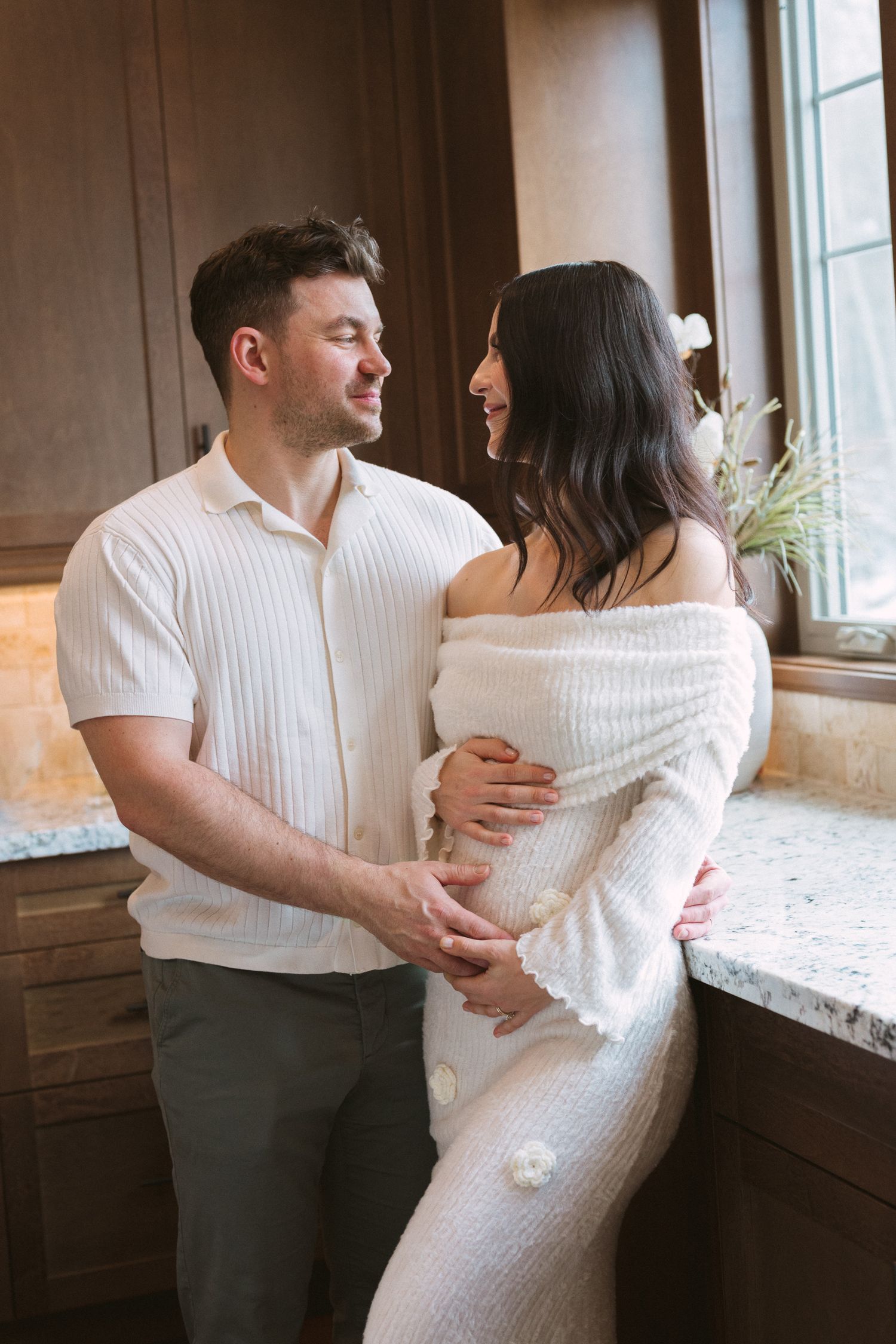 Couple sharing a tender moment in a kitchen, man holding pregnant woman's belly in matching white outfits.