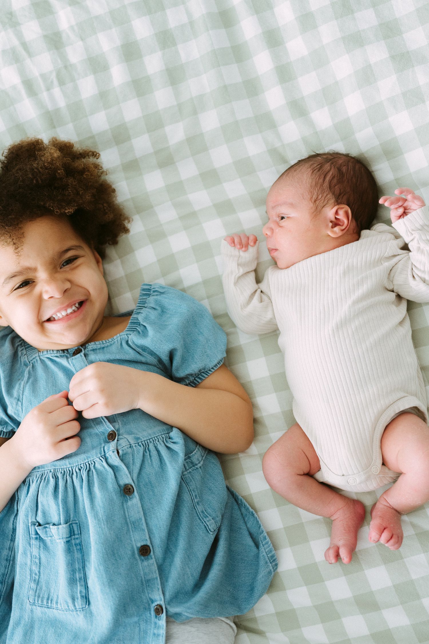 Laughing curly-haired girl in blue denim dress lies next to sleeping newborn on green plaid blanket.