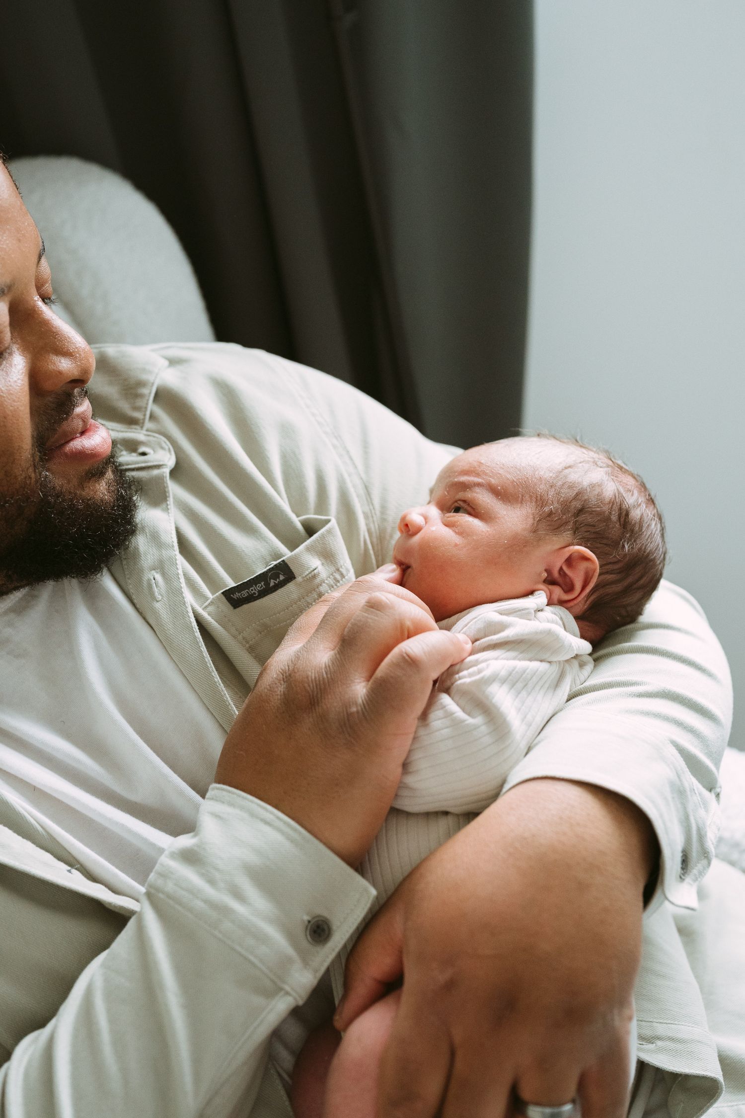 A bearded Black father tenderly cradles his newborn baby in his arms while seated in a chair.