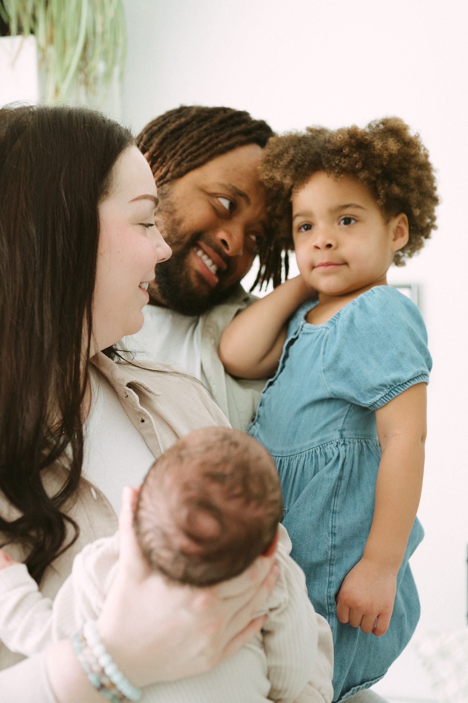Interracial family smiling together, parents holding a newborn and toddler daughter in a bright room.