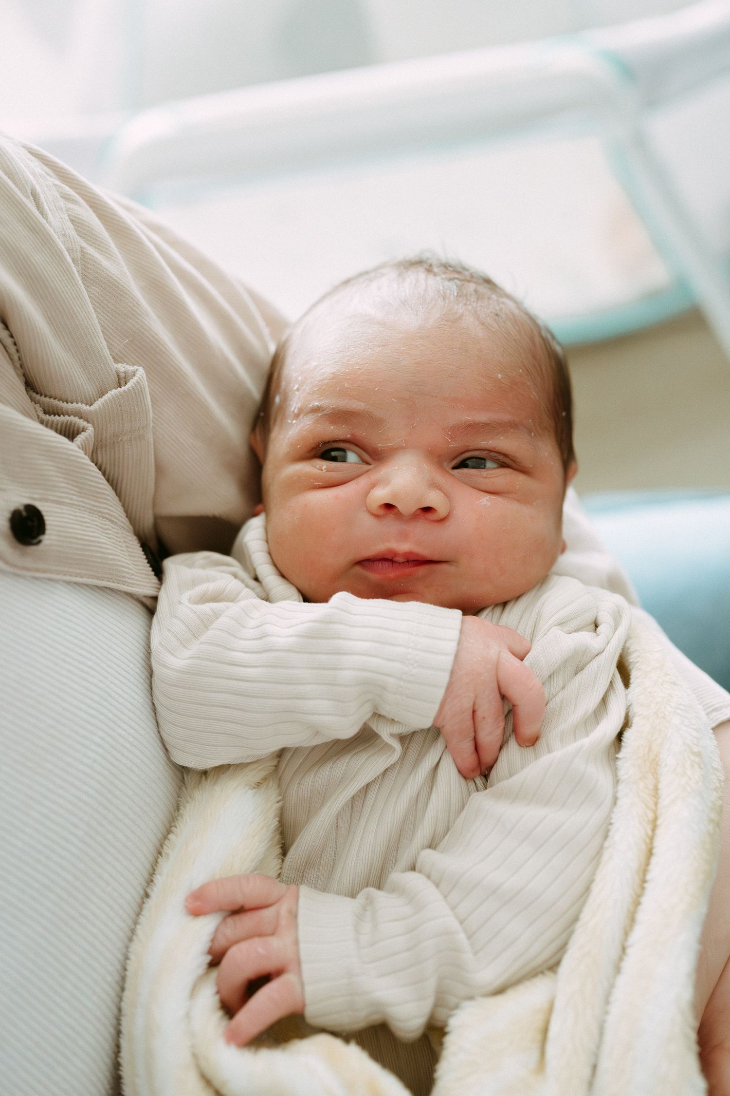 Newborn baby in white outfit cradled in arms, with alert eyes and tiny hands visible against soft blanket.