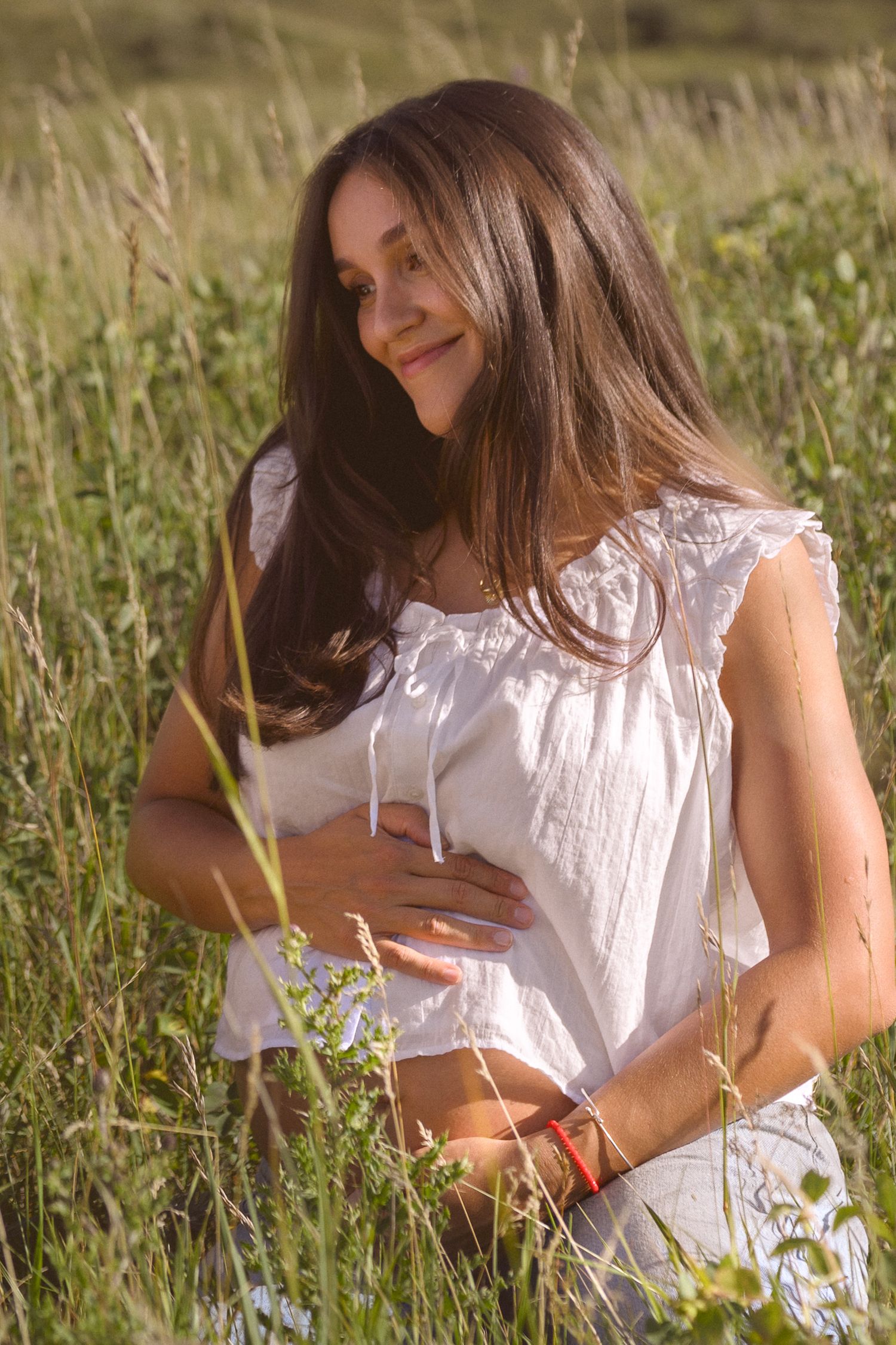 Woman in white blouse sitting among tall grass and wildflowers in golden sunlight.