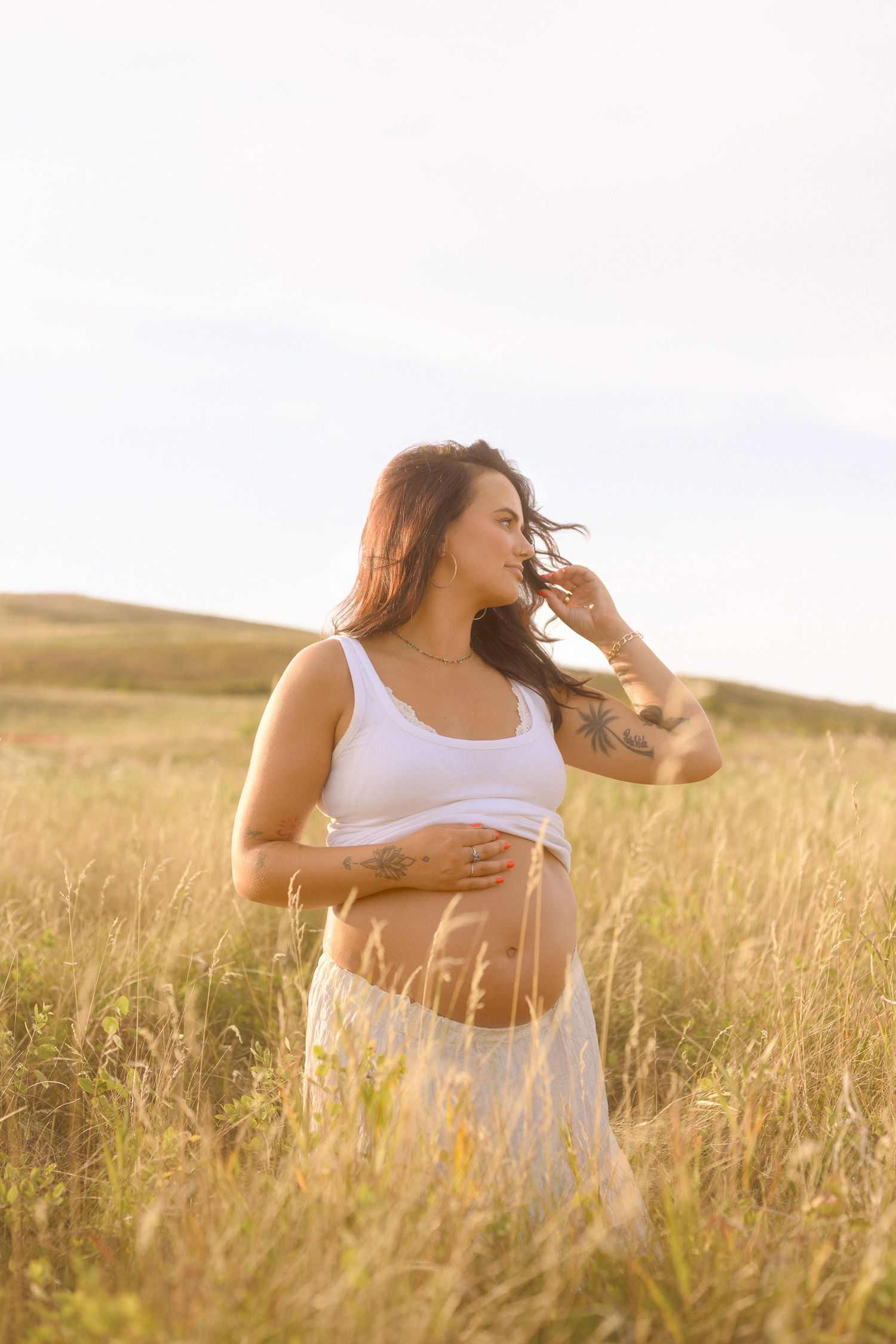 Ethereal maternity portrait in flowing white dress amid tall grass at sunset.