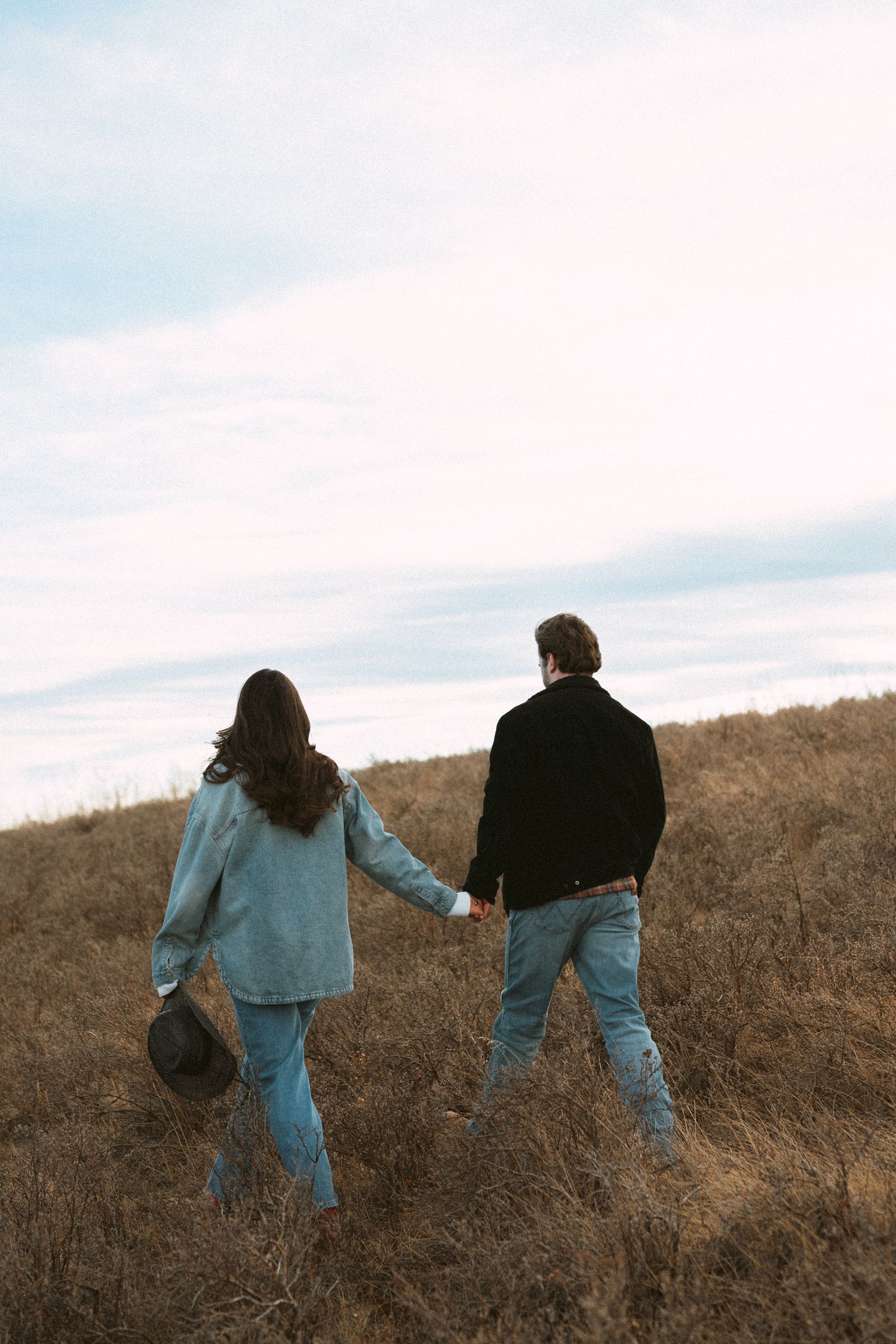 Two people in denim clothing walk together on a brown hillside against a cloudy sky.