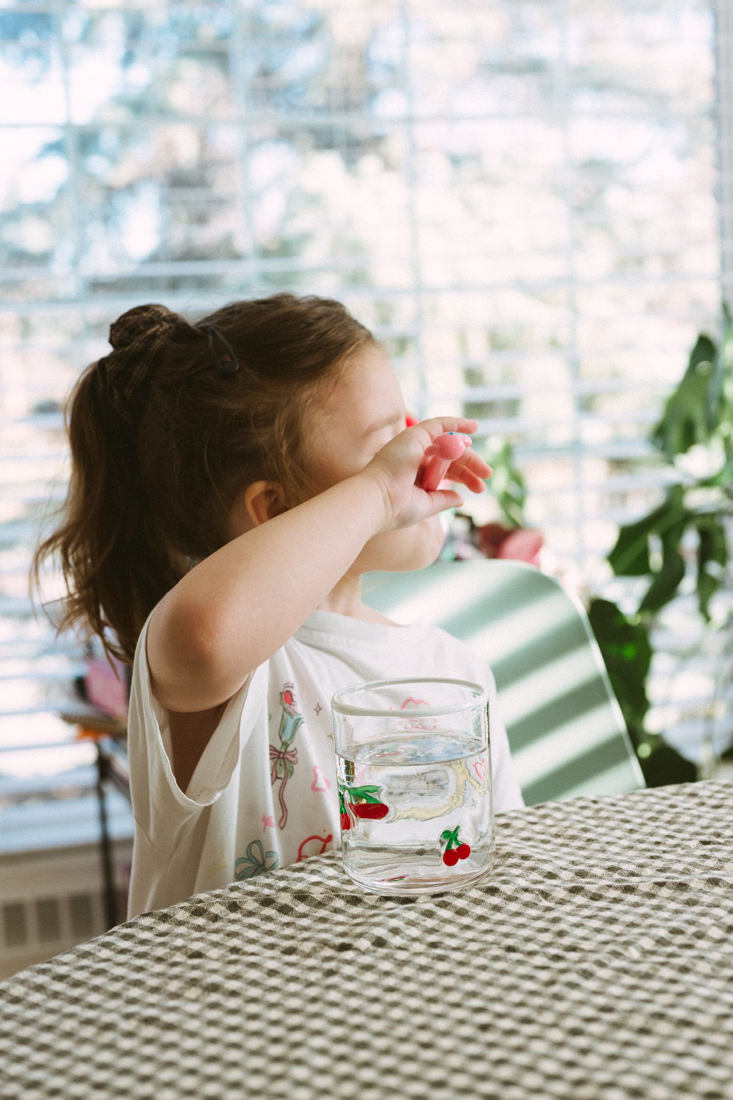 A child drinks from a glass with a checkered tablecloth and window light in the background.